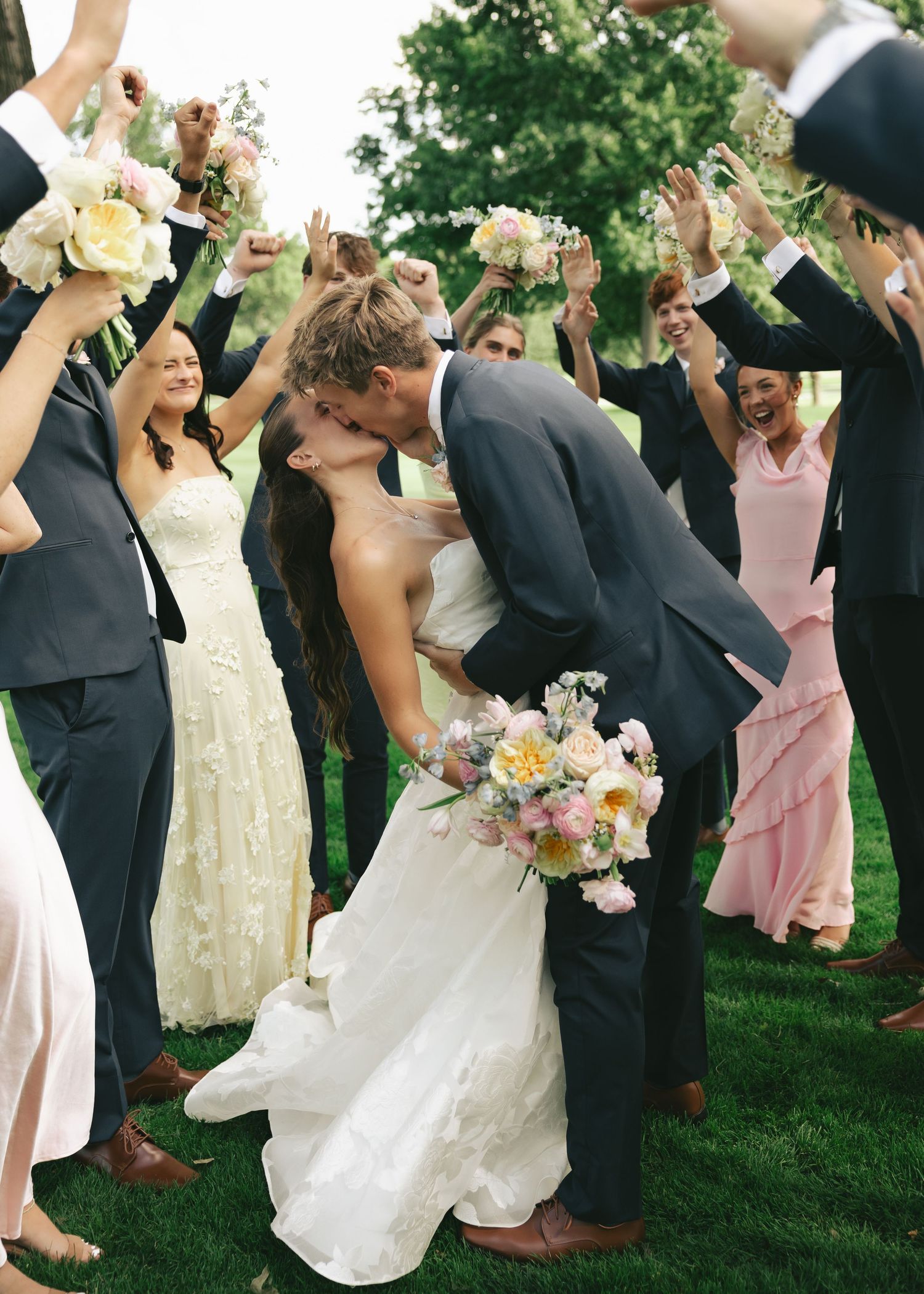 Bride and groom share a romantic kiss surrounded by cheering wedding party on a sunny outdoor lawn.