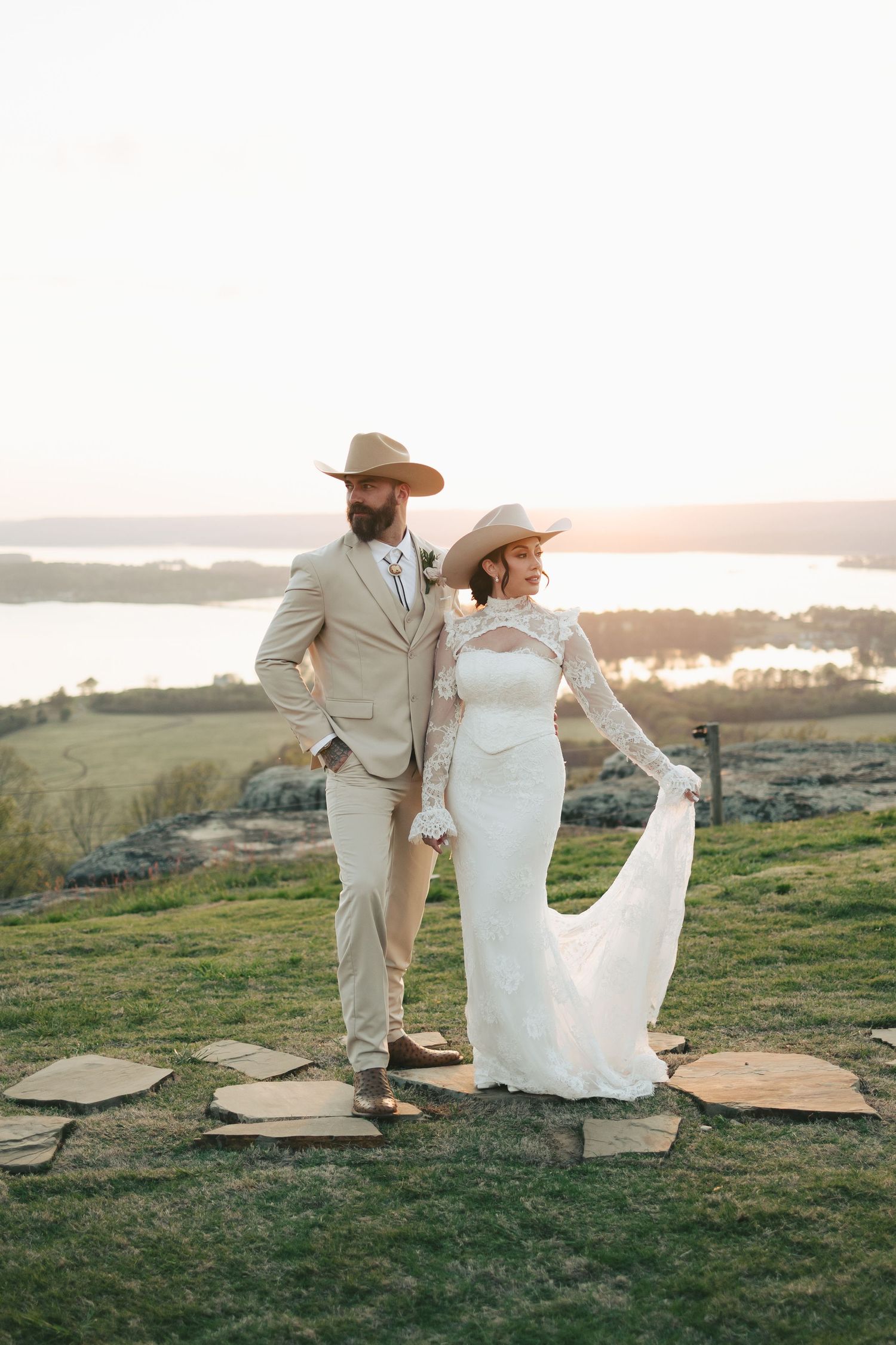 Bride and groom in cowboy hats pose at golden hour with scenic lake views behind them.