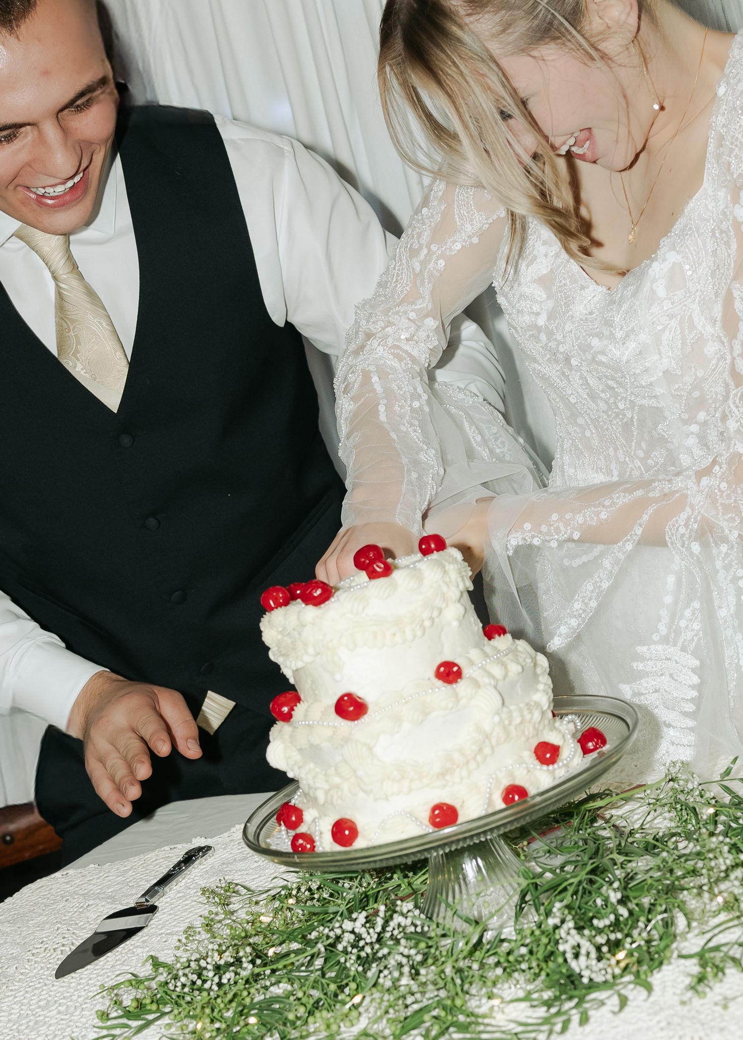 Smiling bride and groom cut a two-tier white wedding cake decorated with red cherries at reception.