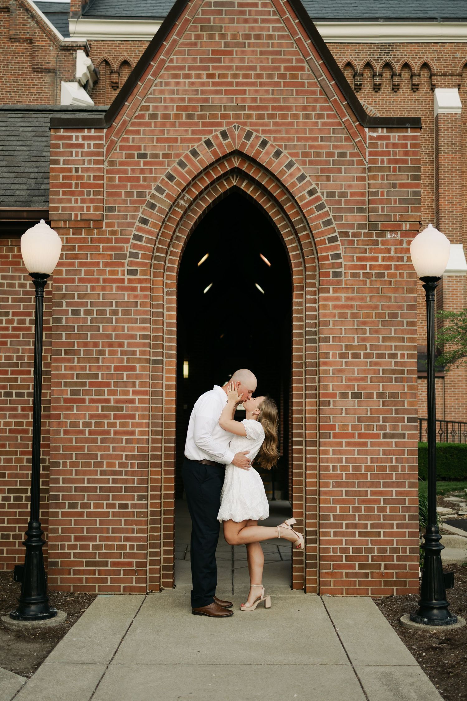 Couple sharing romantic kiss in Gothic brick church doorway, woman in white mini dress lifting one leg.