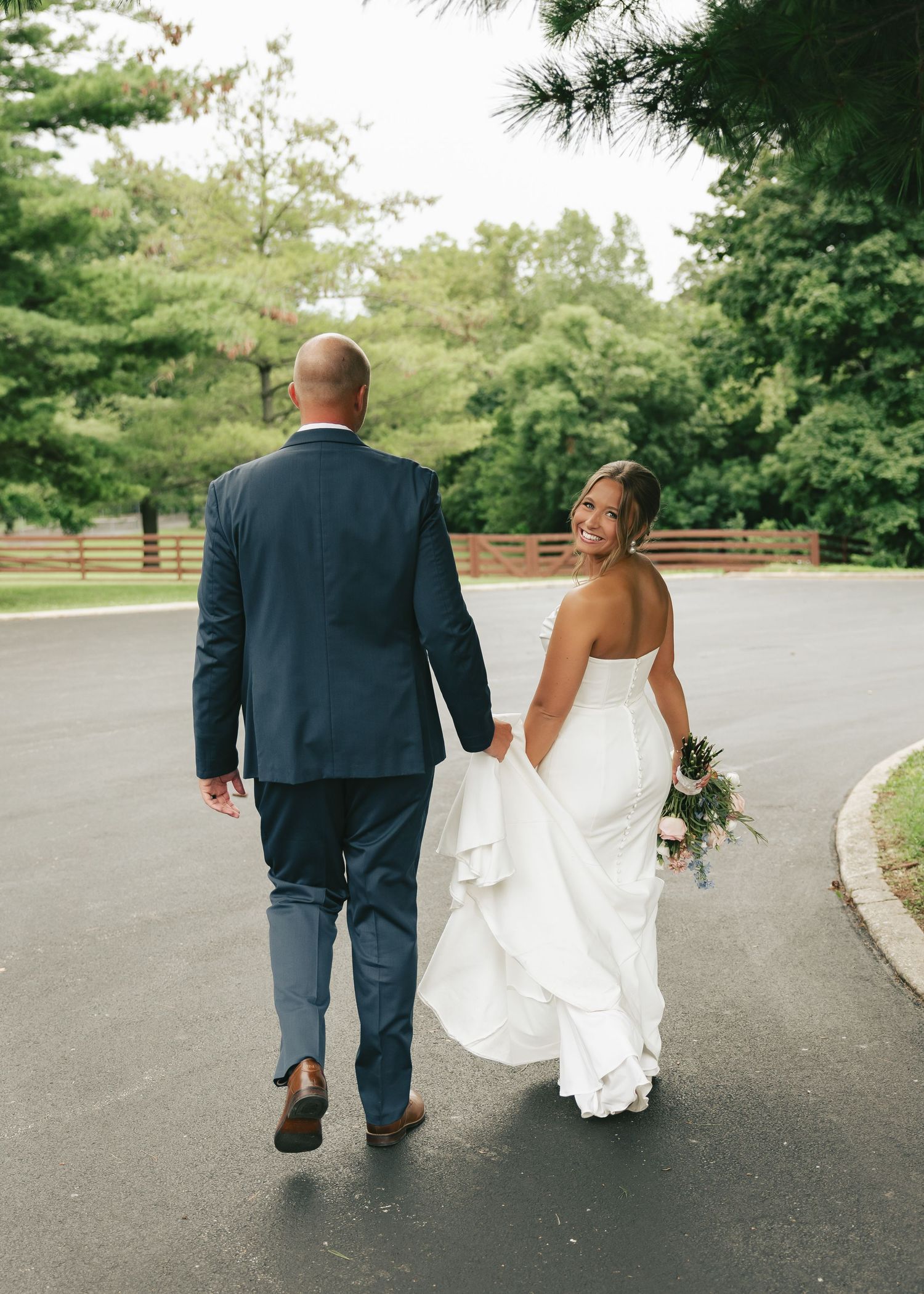 Bride and groom walking hand-in-hand down a paved path, bride in white gown holding bouquet, groom in navy suit.