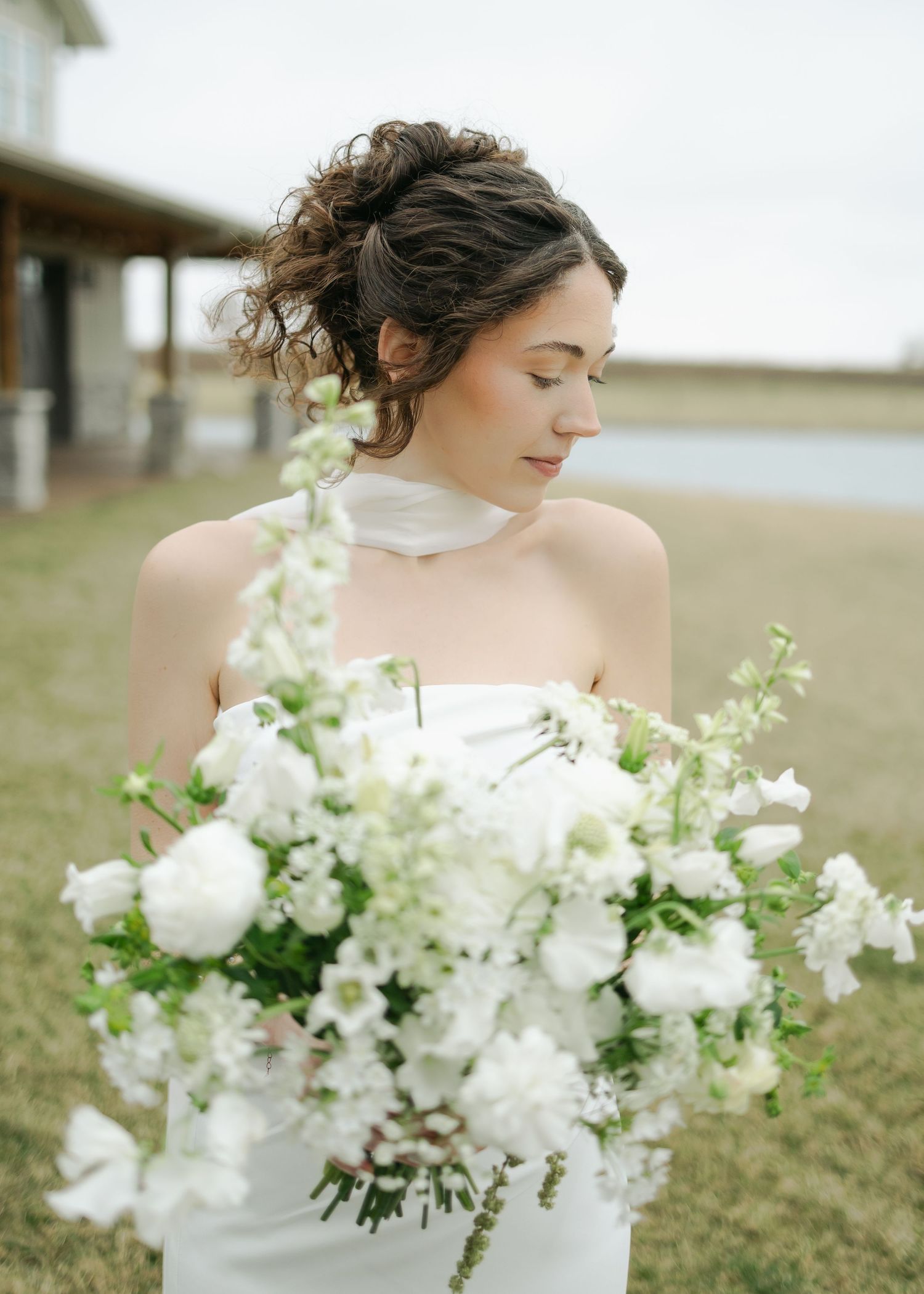 Bride with updo holds large white floral bouquet with eyes closed near waterfront in white gown.