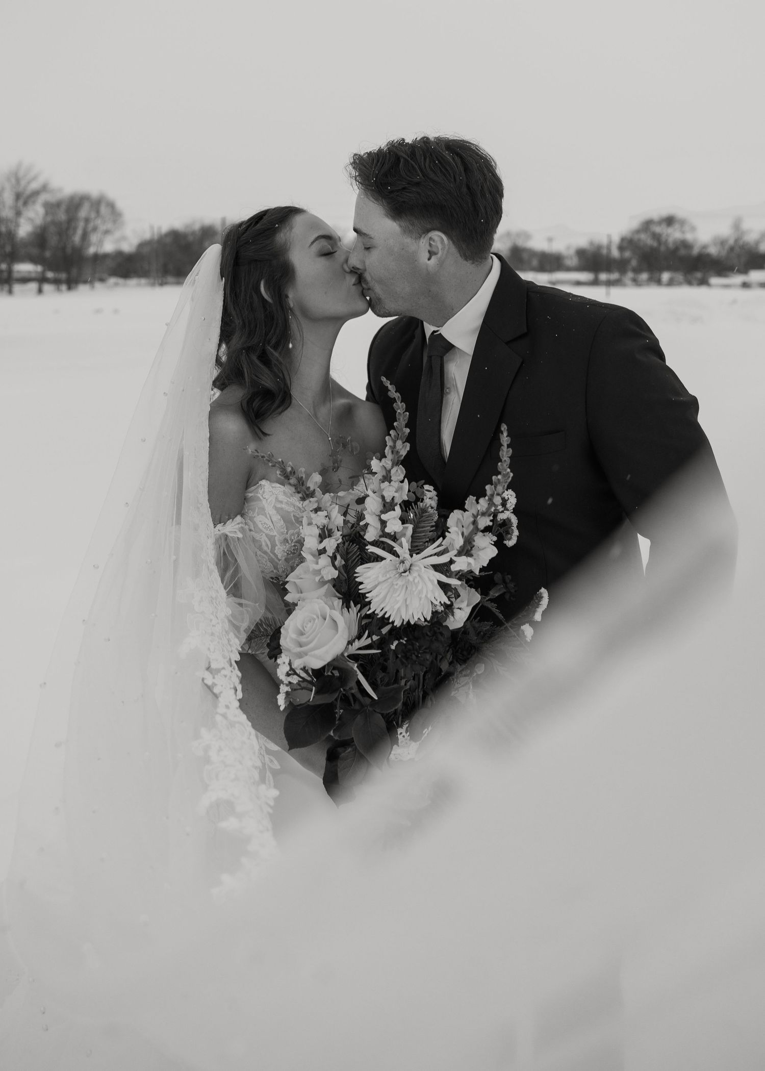 Black and white winter wedding photo of a bride and groom kissing in a snowy landscape with veil.