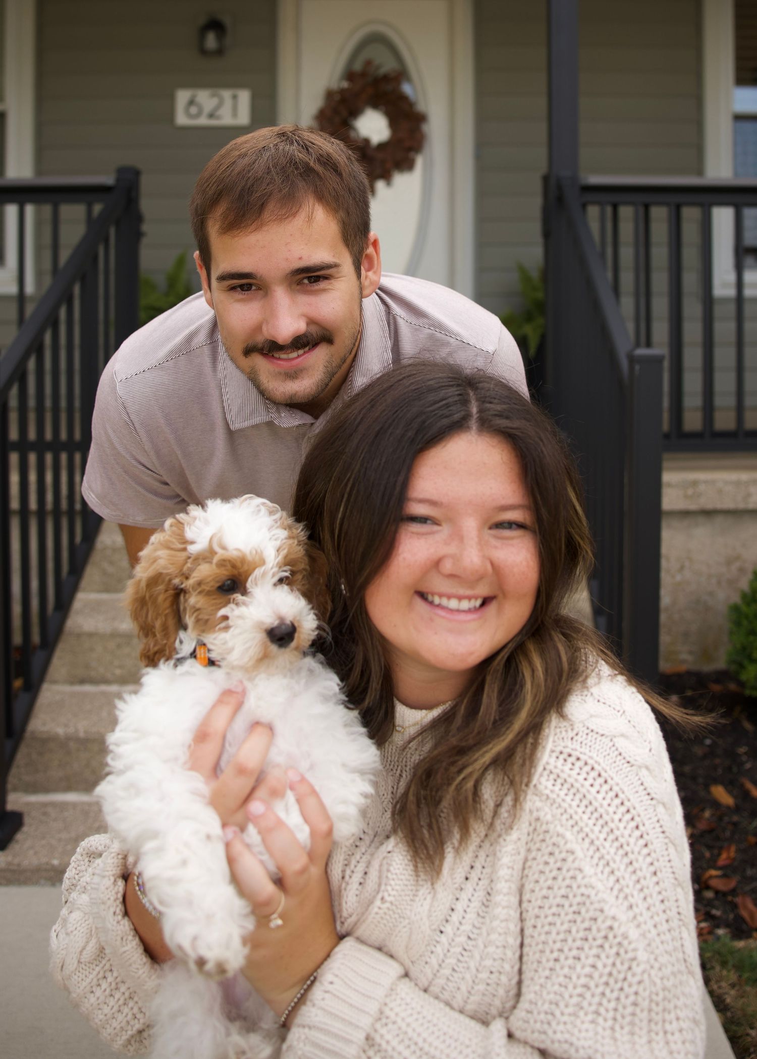 Smiling couple posing with an adorable Cavapoo puppy in front of their home at address 621.