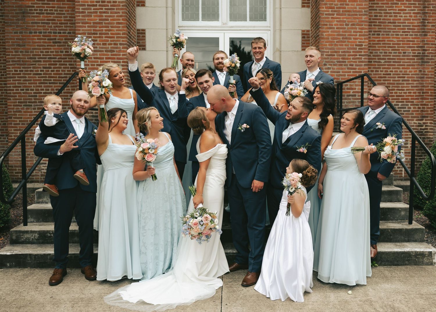 Wedding party celebrating with bride and groom kissing, bridesmaids in light blue dresses, groomsmen in navy suits.