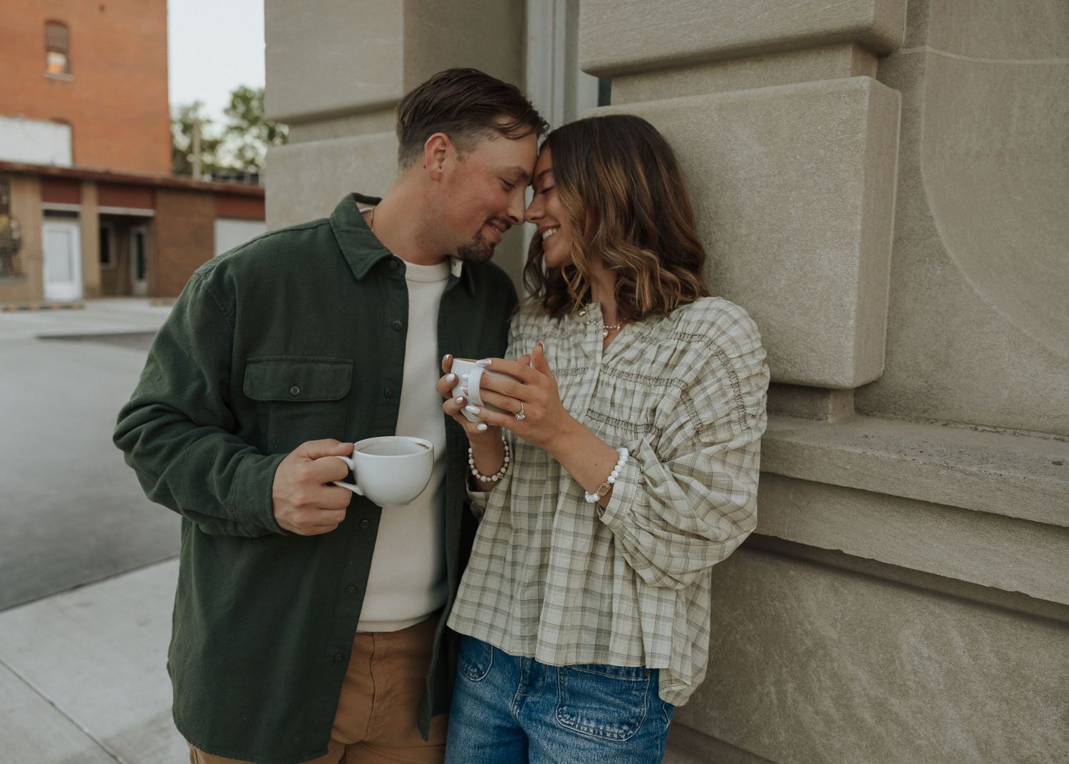 Couple sharing a romantic moment with coffee cups leaning against a stone building.