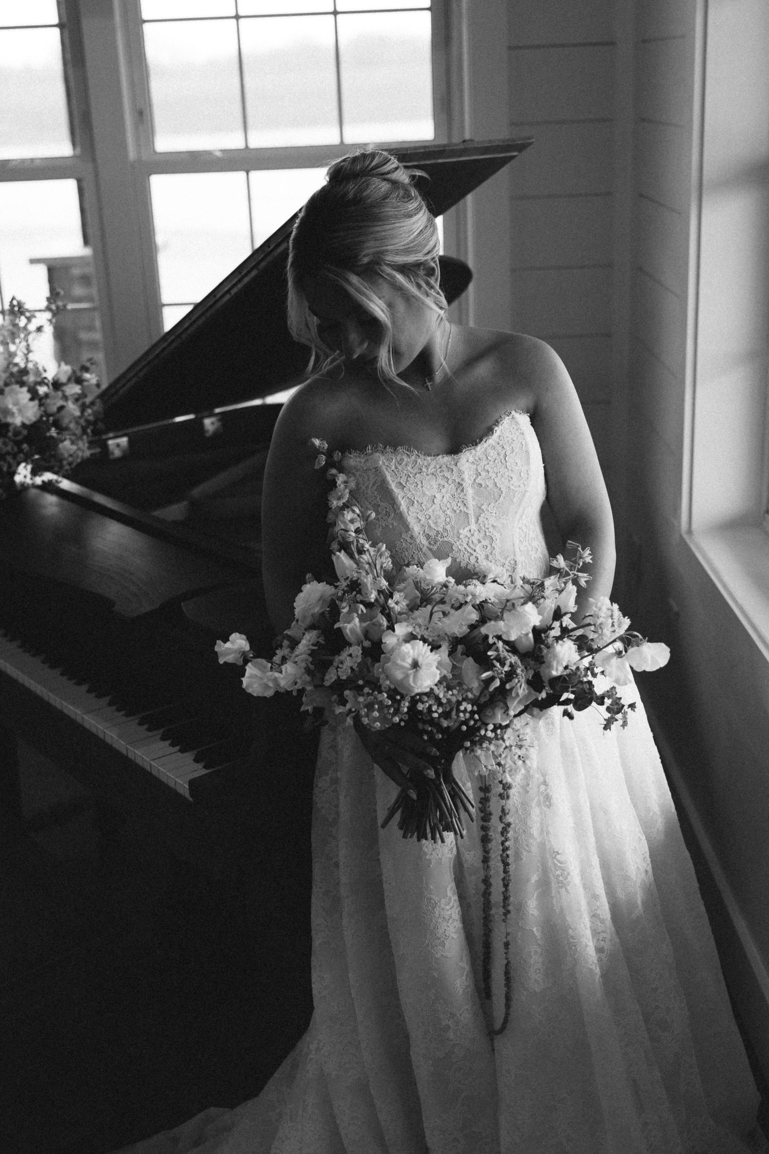 Bride in strapless lace gown holding bouquet beside grand piano in black and white photo.