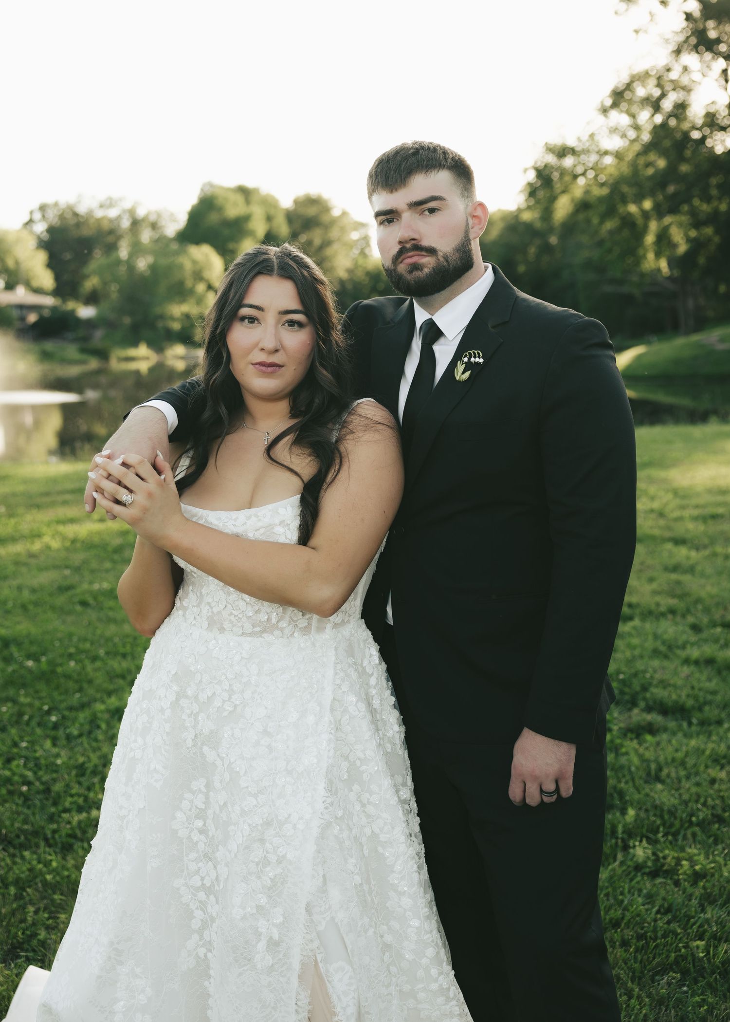 Bride in white lace gown and groom in black suit pose together outdoors at a wedding ceremony.