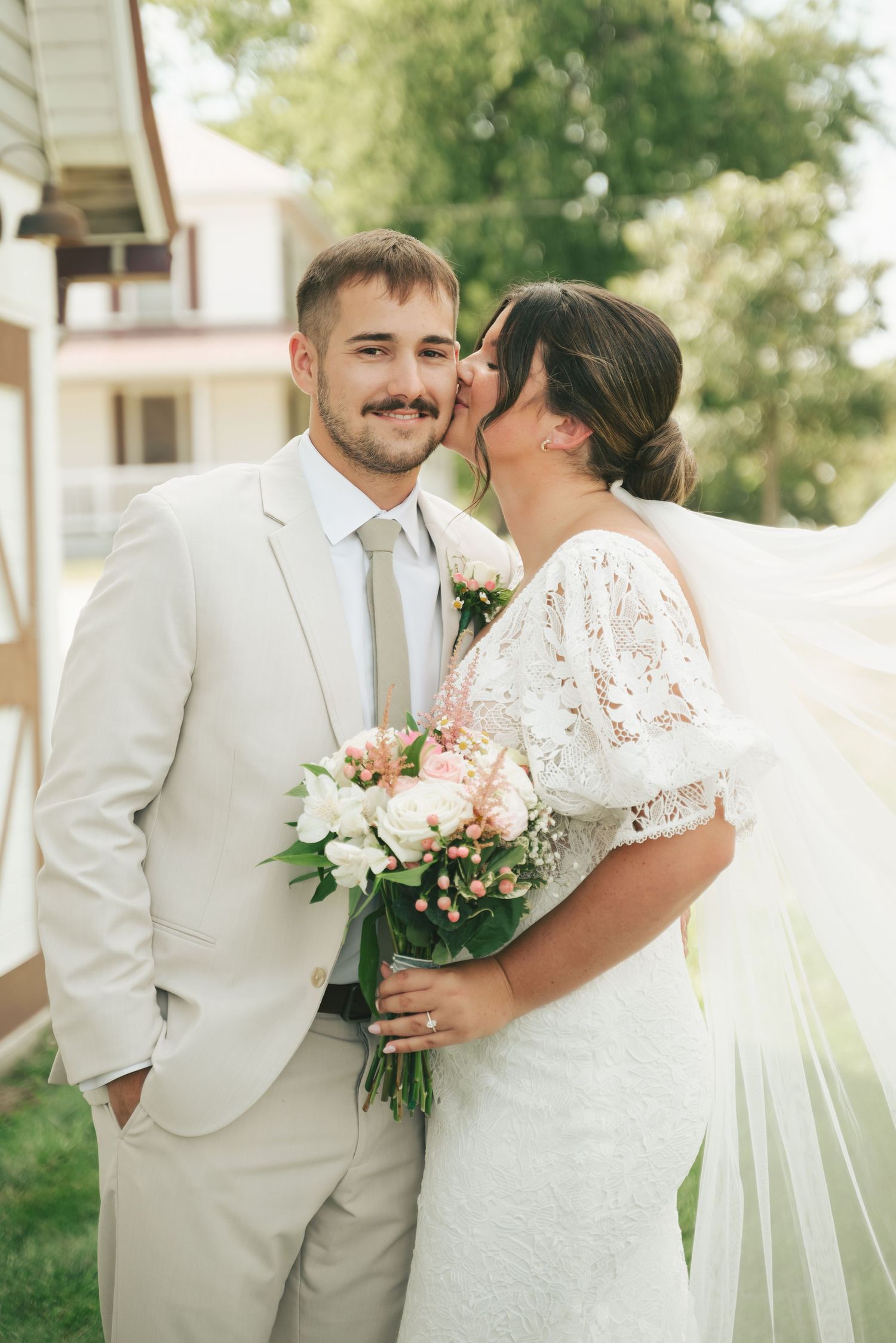 Bride in lace gown kisses groom in tan suit holding pink and white bouquet outdoors at summer wedding.