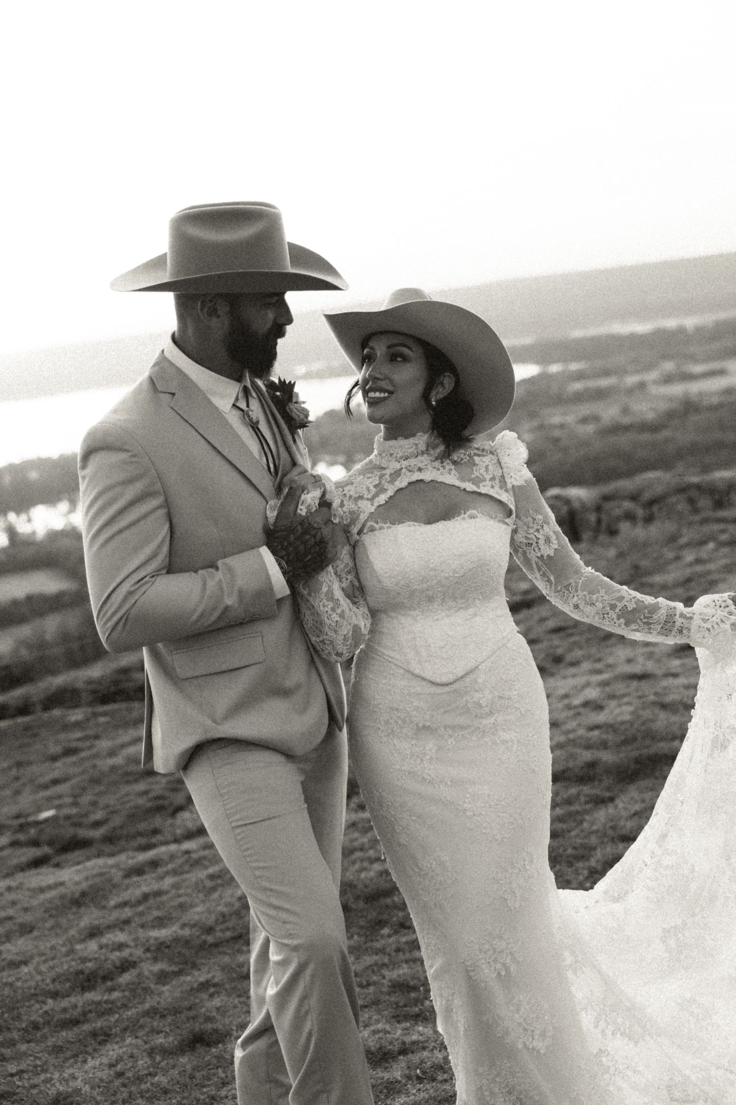 Stylish bride and groom in cowboy hats posing outdoors at sunset in black and white wedding photo.