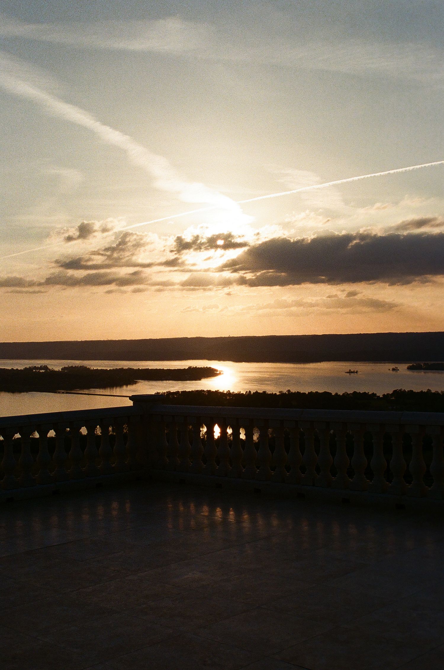 Golden sunset over a tranquil lake viewed from an elegant balustrade terrace with dramatic clouds and sun rays.