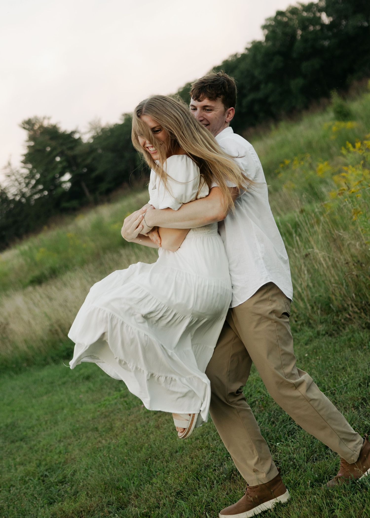 Couple laughing and dancing in a green field, man lifting woman in white maxi dress during outdoor engagement session.