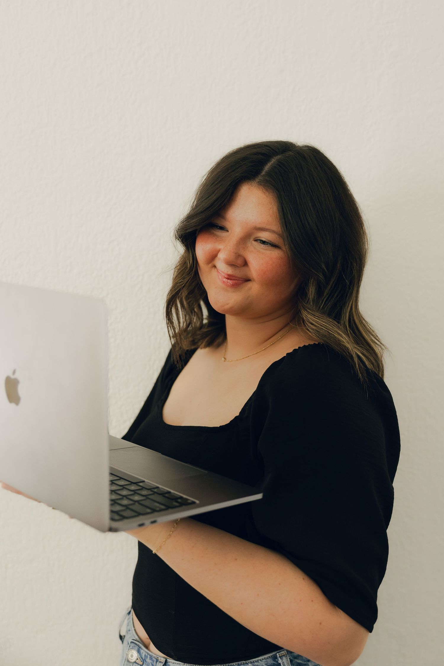 Young woman smiling while holding an Apple MacBook laptop against a white wall.