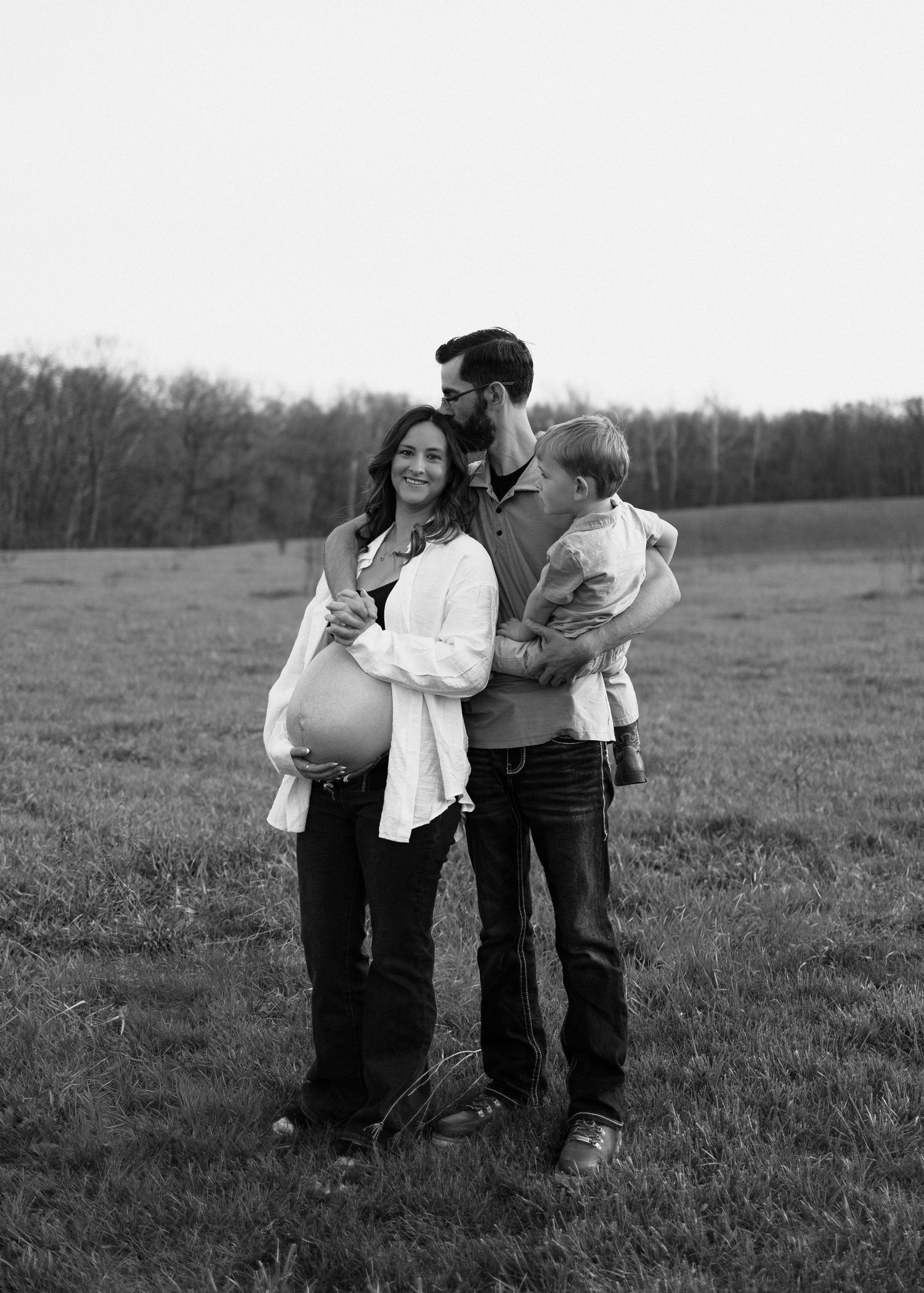Black and white family portrait of pregnant woman, man, and toddler standing in an open field.