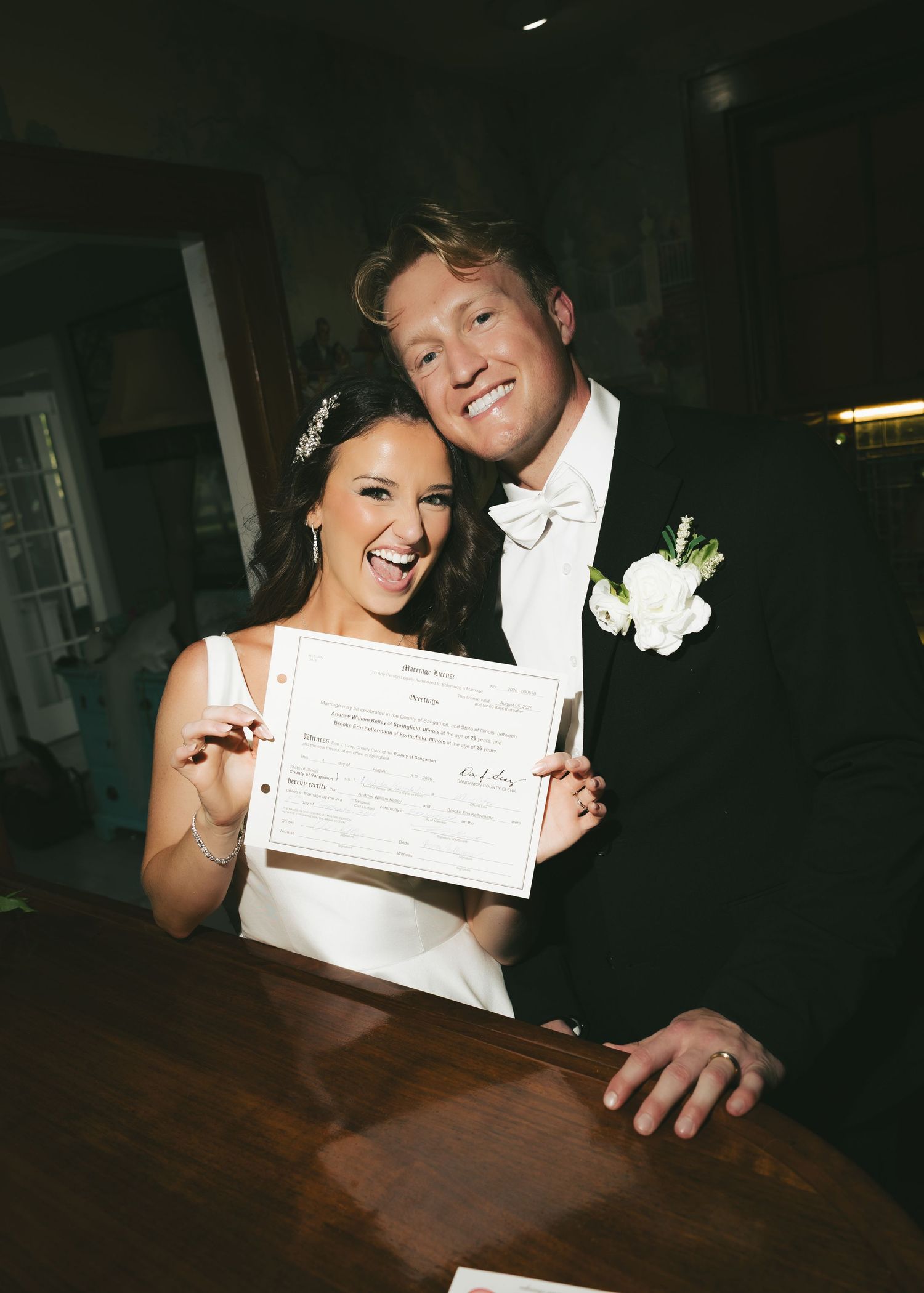 Happy newlyweds smiling and holding their marriage certificate, bride in white gown and groom in black tuxedo with white bow tie.