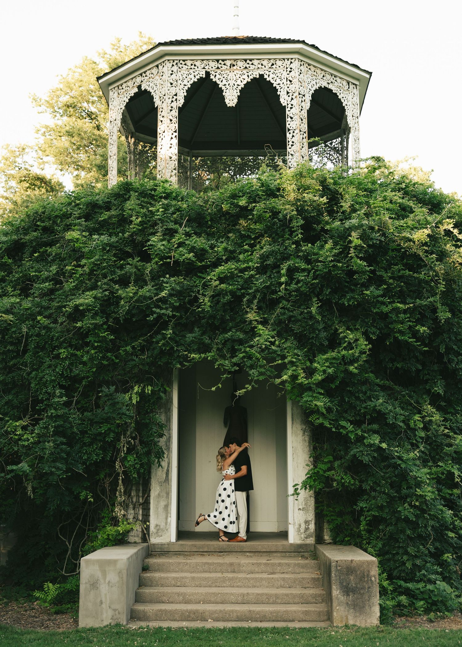 Couple embracing in ivy-covered gazebo with ornate white lattice architecture and stone steps in a lush garden setting.