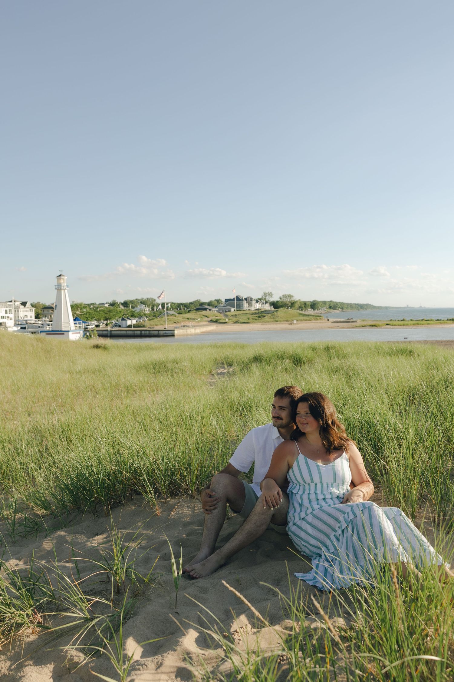 Couple sitting together on sandy beach dunes with green grass, lighthouse, and calm waterway in background on sunny day.