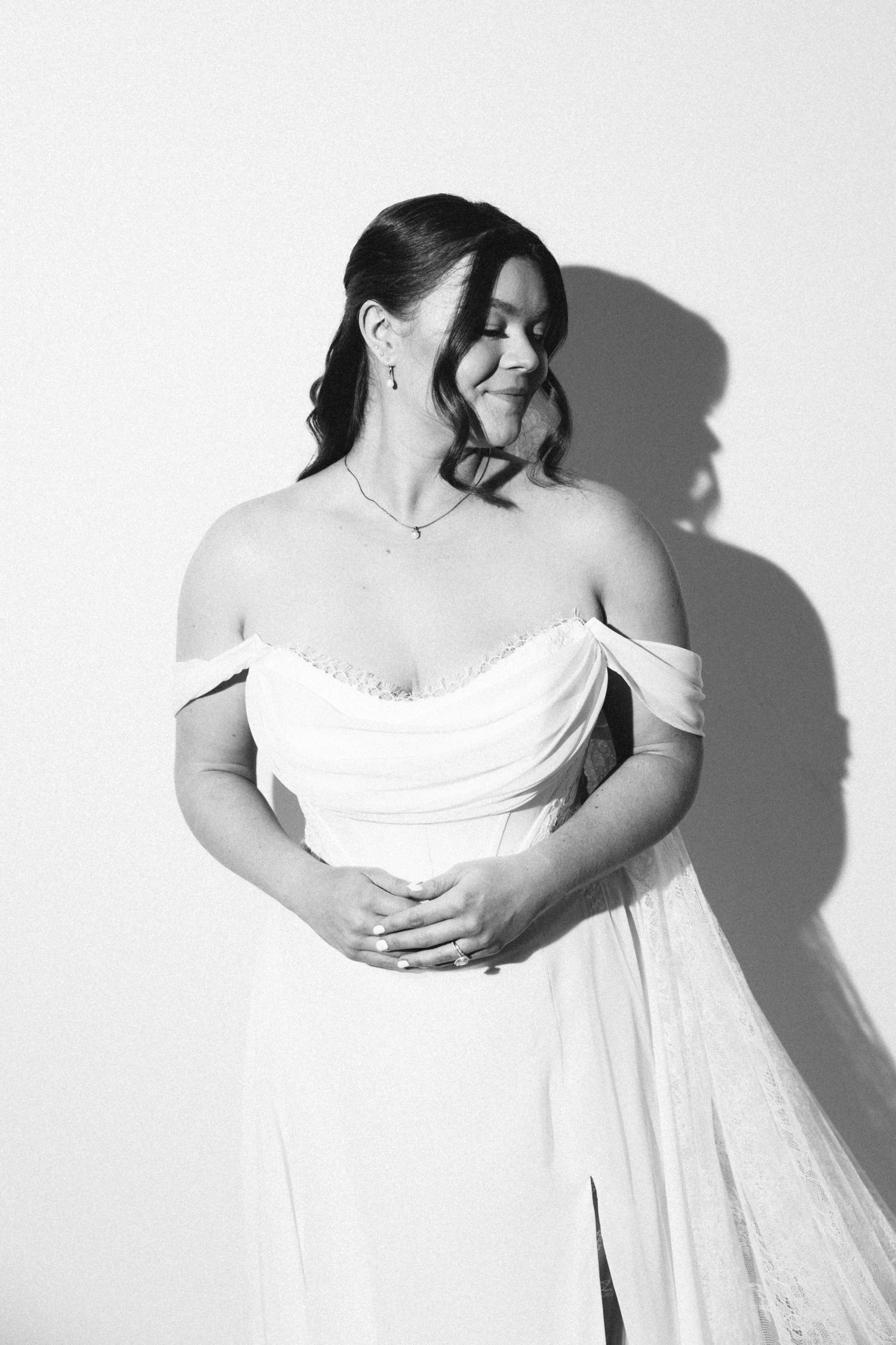 Black and white portrait of bride in off-shoulder draped wedding gown smiling softly against white wall.