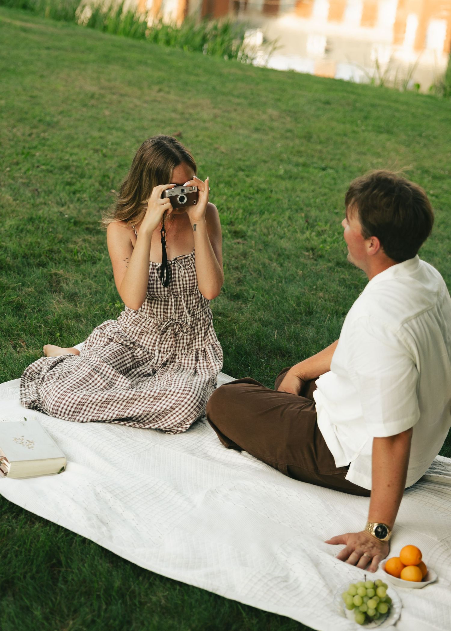 Couple enjoying a picnic on a blanket in the grass, woman photographing man with vintage camera.