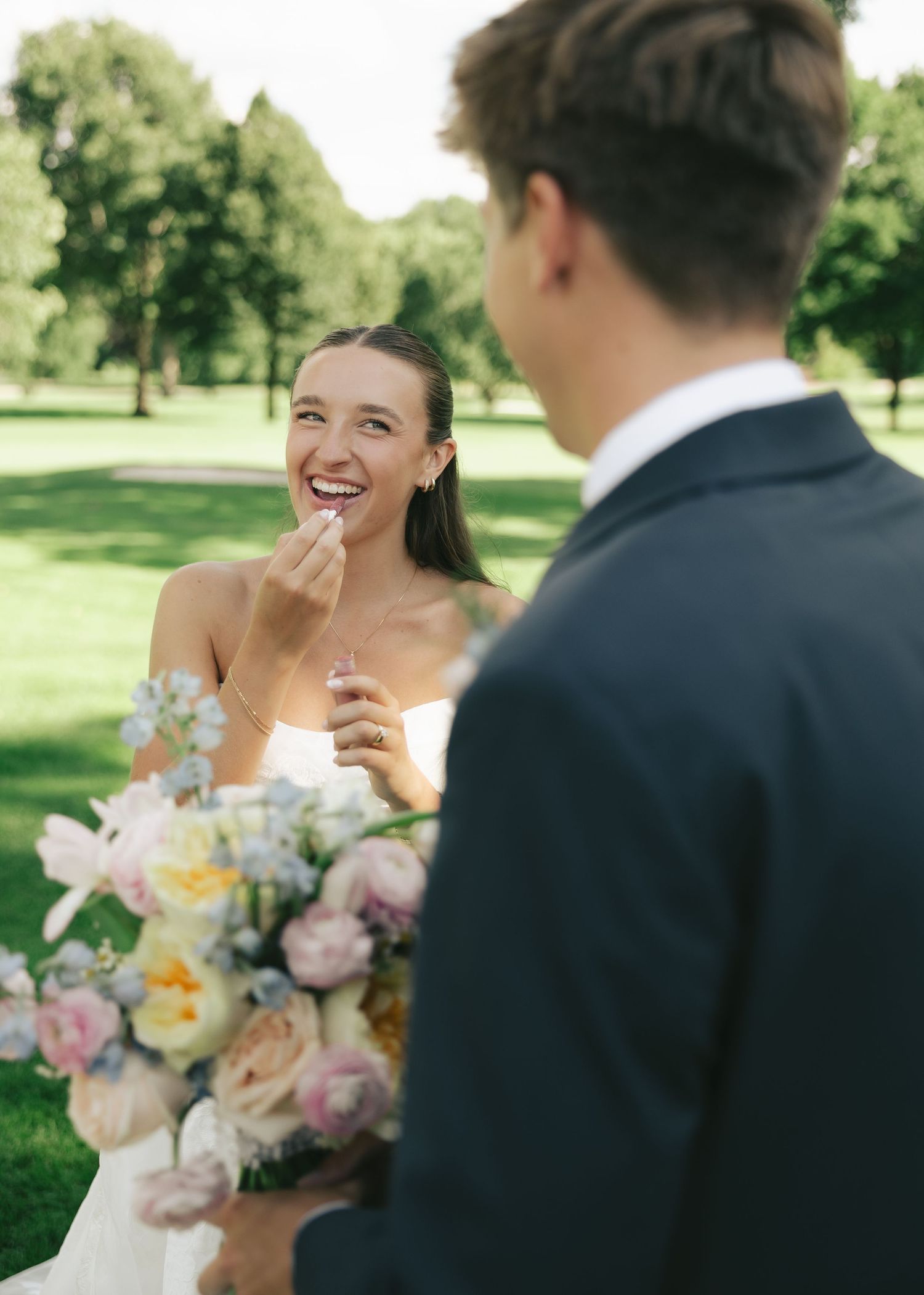Smiling bride holding pastel bouquet laughs with groom in navy suit during outdoor wedding ceremony on lush green grounds.