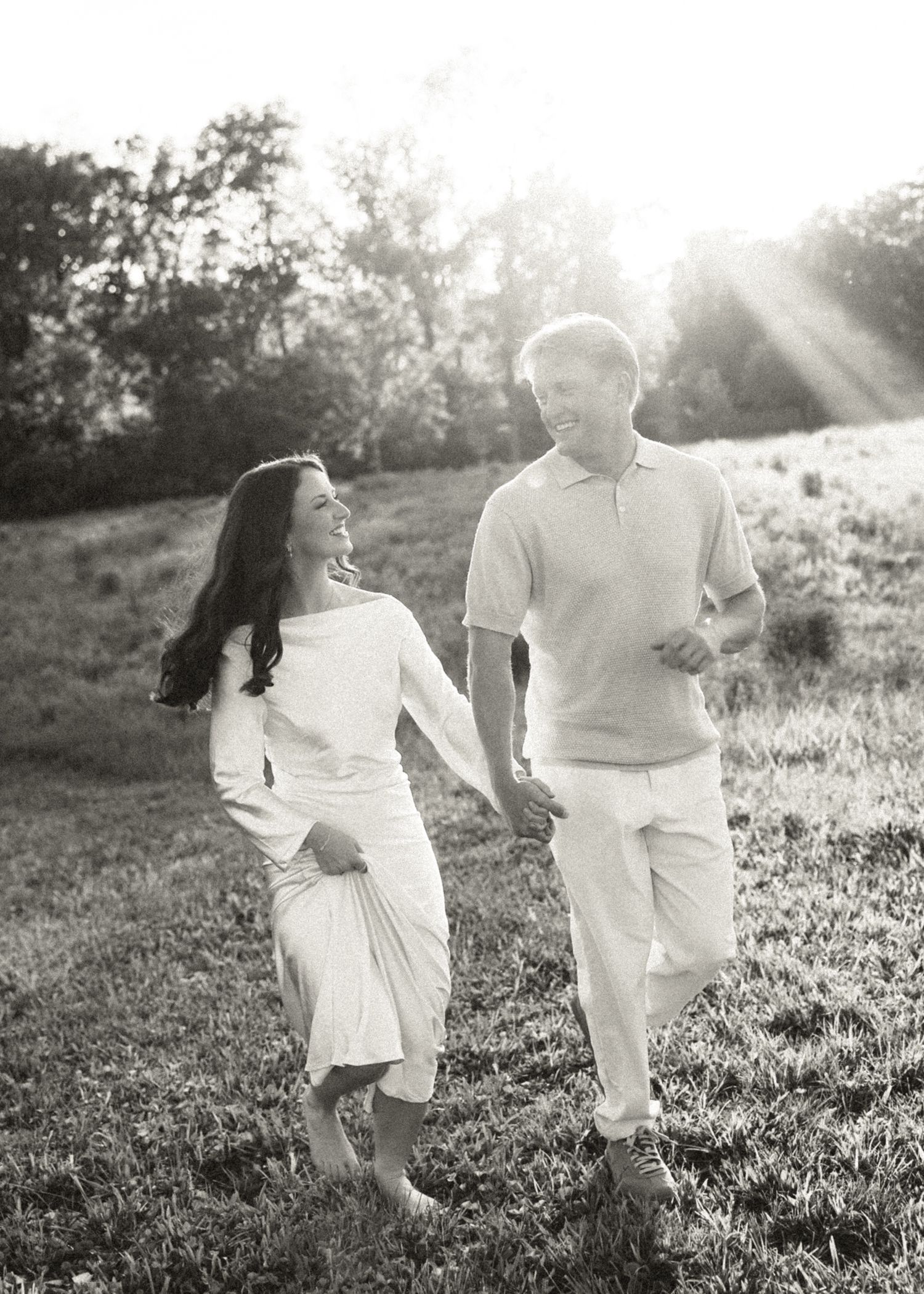 Black and white photo of a smiling couple holding hands and walking through a sunlit grassy field.