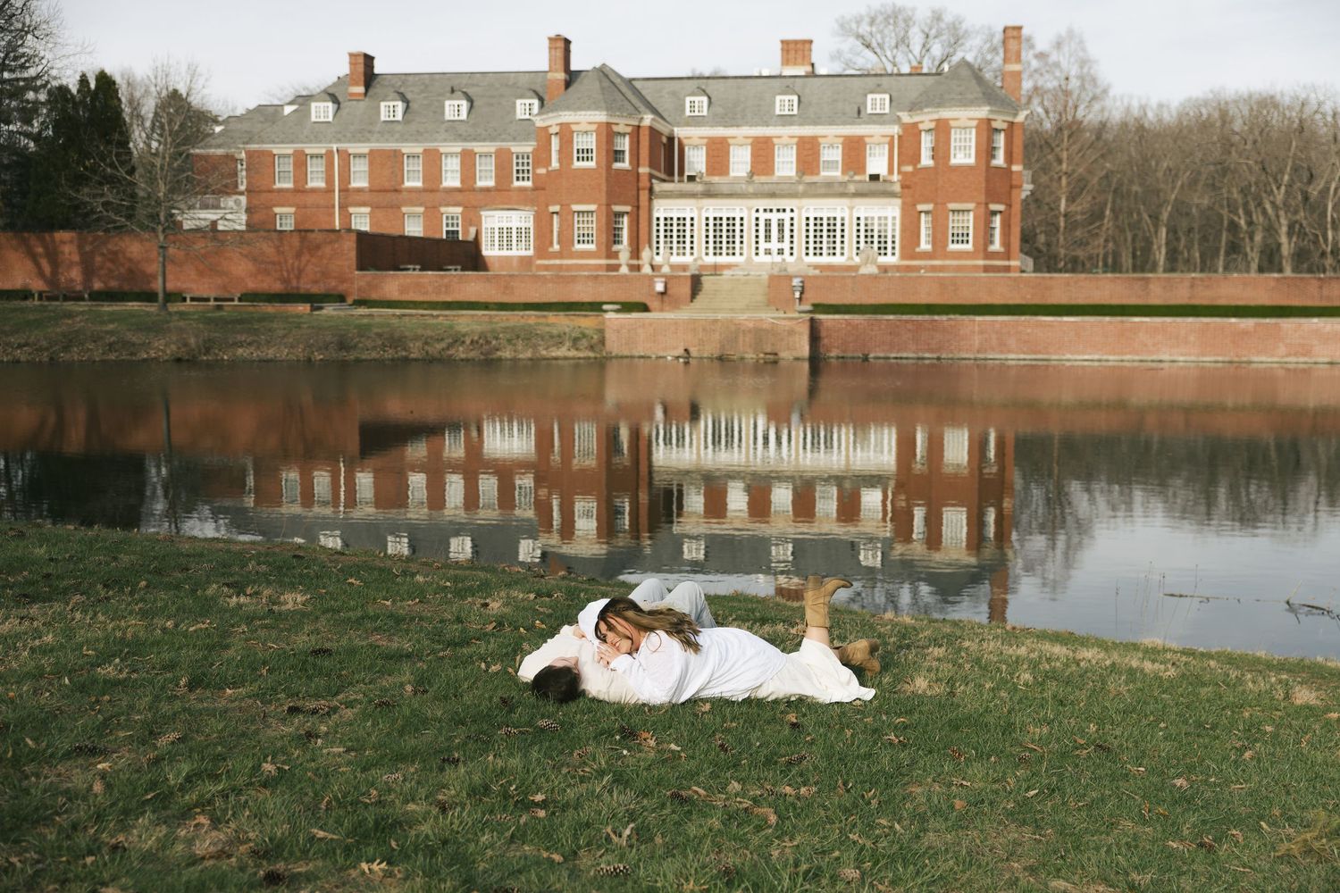 Couple in wedding attire lying on grass beside a reflective pond with Allerton Park grand estate behind them.