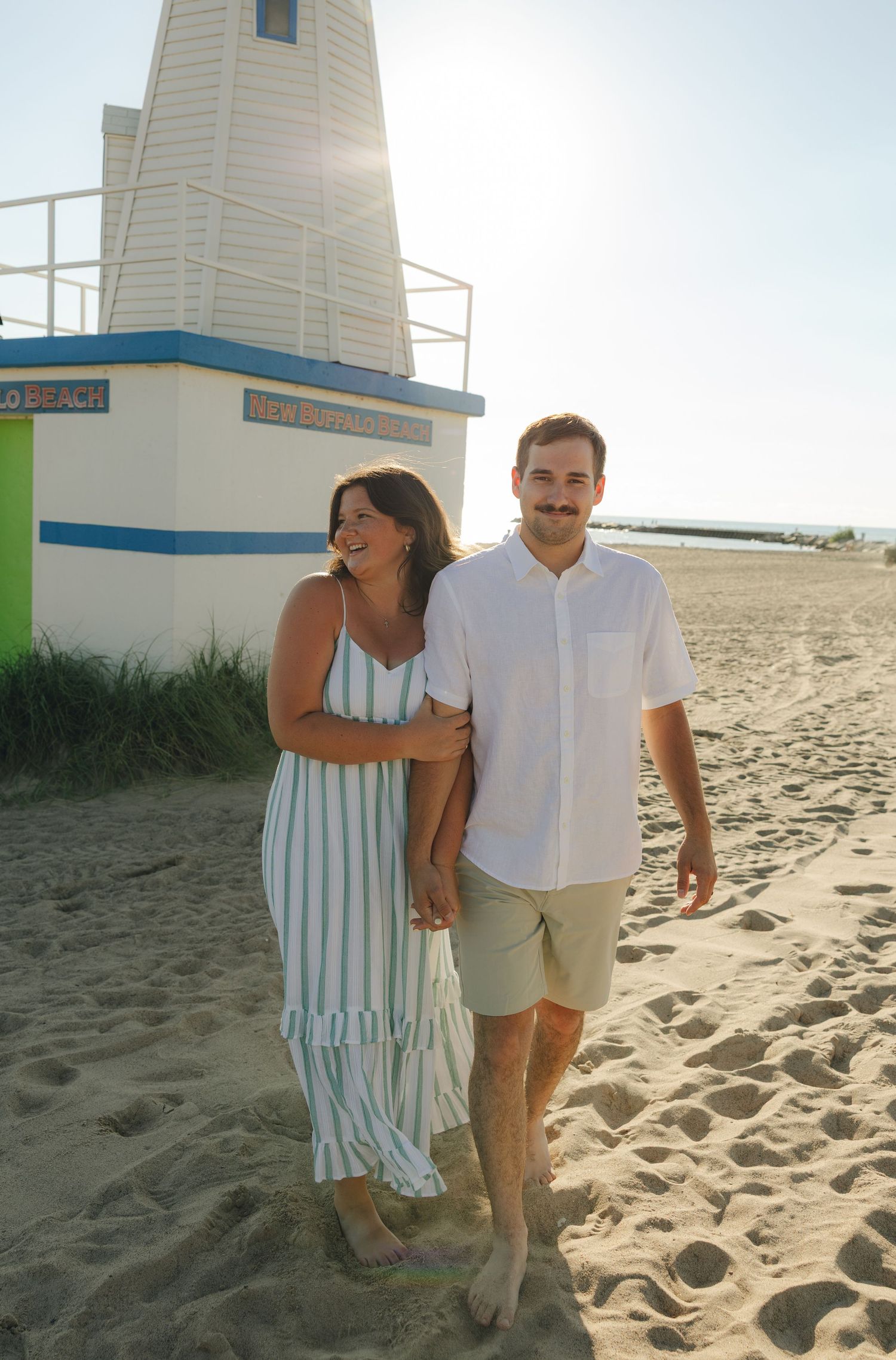 Couple walking hand in hand on sandy beach near white lighthouse, woman in striped dress, man in white shirt and khaki shorts.