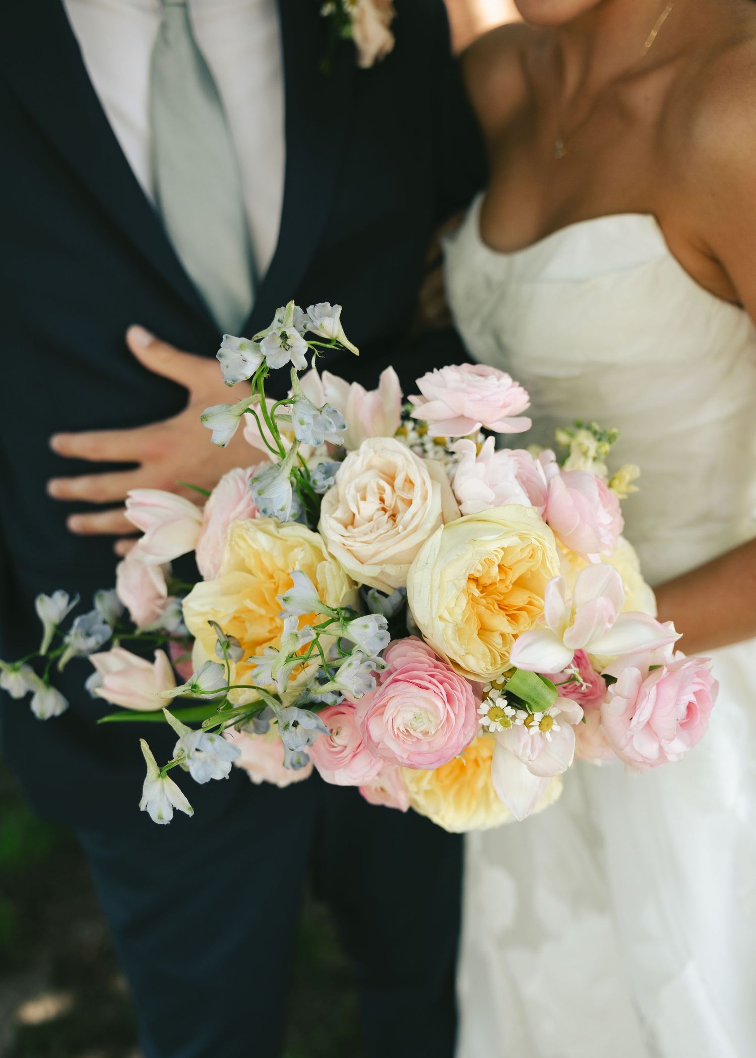 Bride holding a pastel wedding bouquet with pink roses, yellow garden roses, and blue delphinium beside groom.