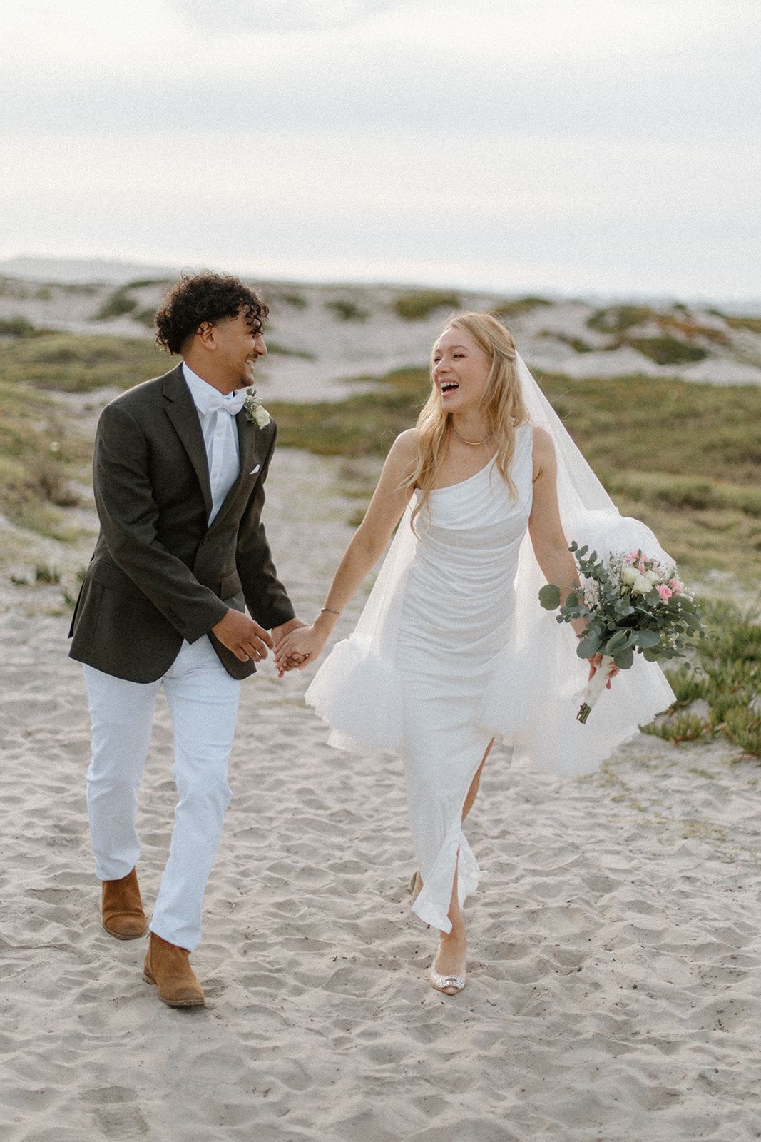 A couple in wedding attire playfully walks and dances together on a sandy beach during their coastal celebration.