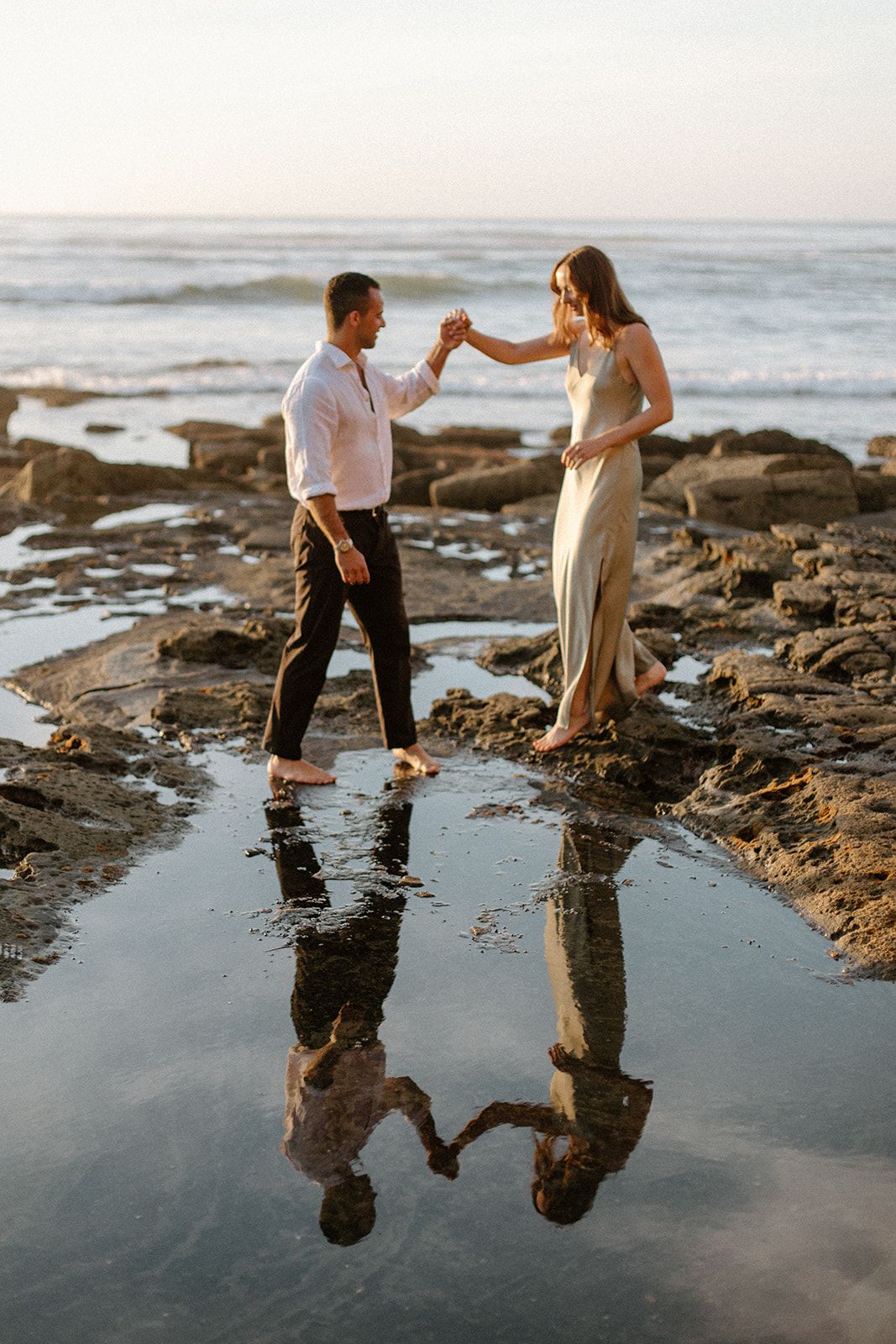 A couple walks along a rocky beach at sunset with their reflections mirrored in the tidal pools.