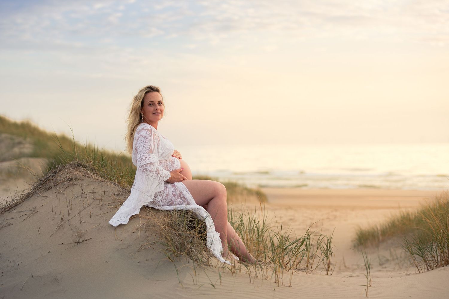 Pregnant woman in white lace dress sitting on sand dunes at beach during golden hour sunset.