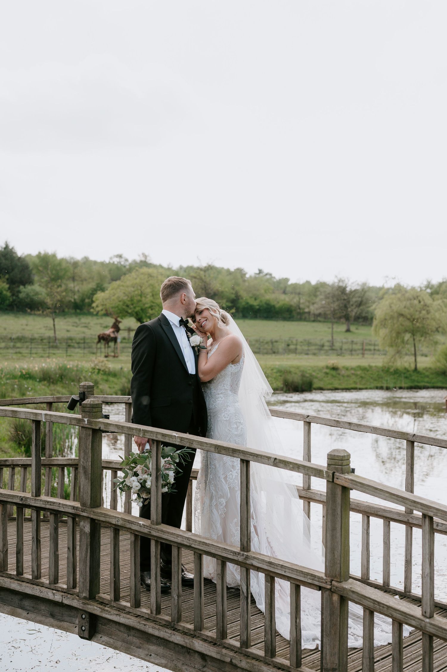 A couple shares an intimate moment on a rustic wooden bridge overlooking a pastoral countryside setting.