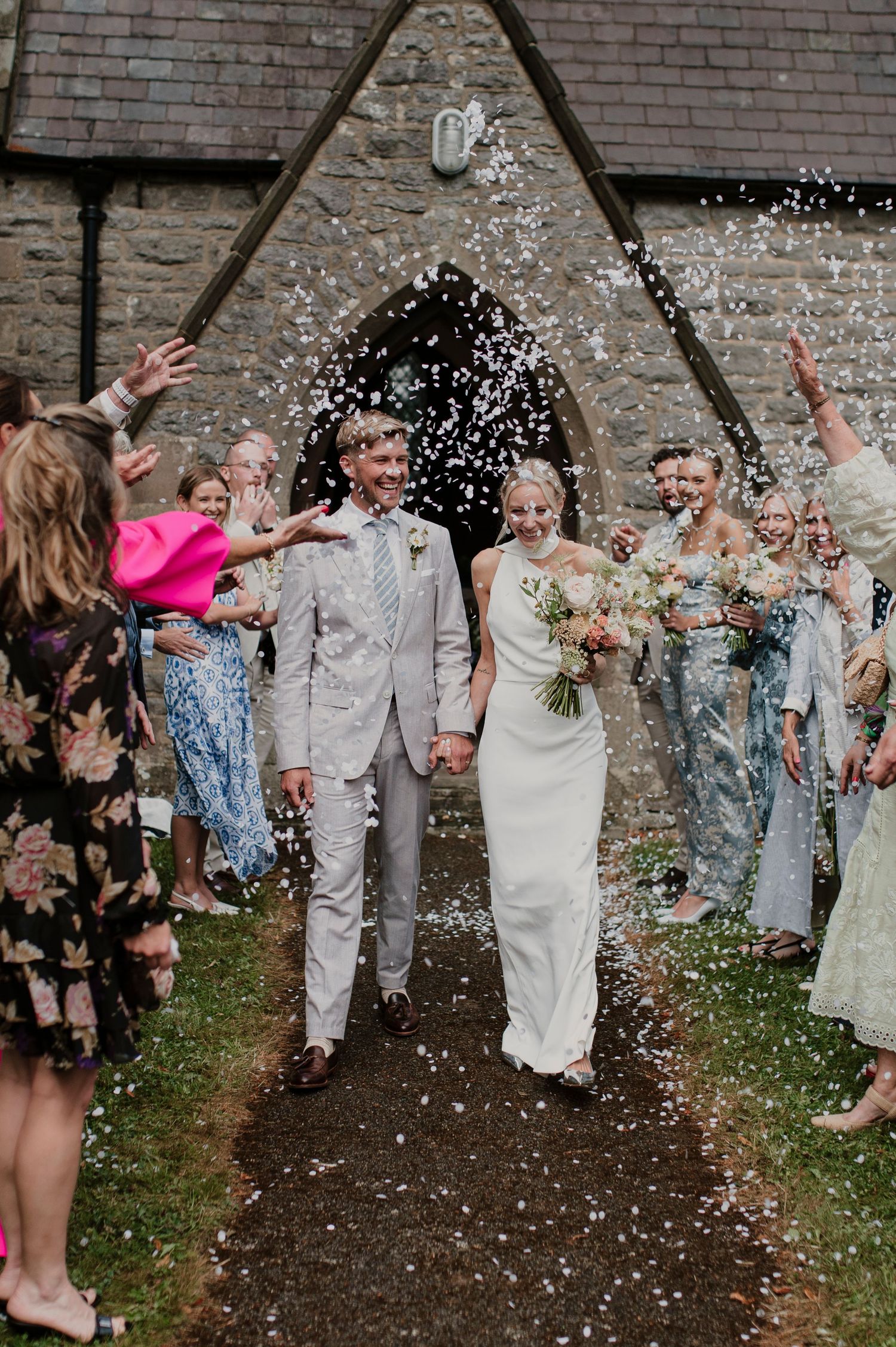 Wedding guests shower the newlyweds with white confetti outside a brick building.