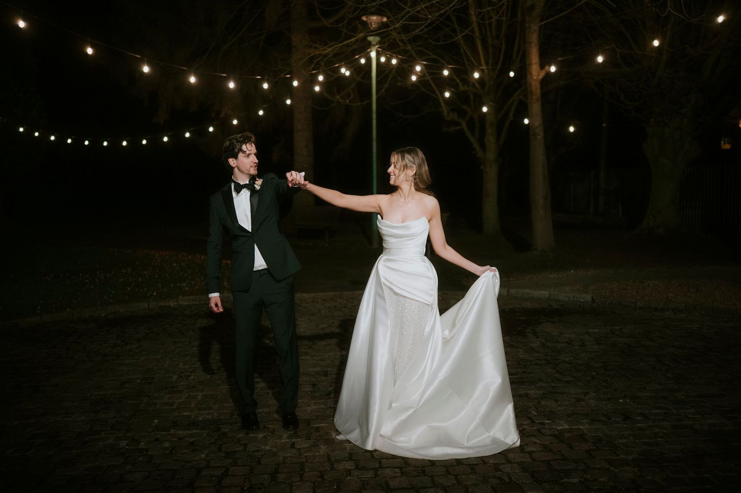 A couple dances together at night under string lights in an outdoor wedding reception setting.