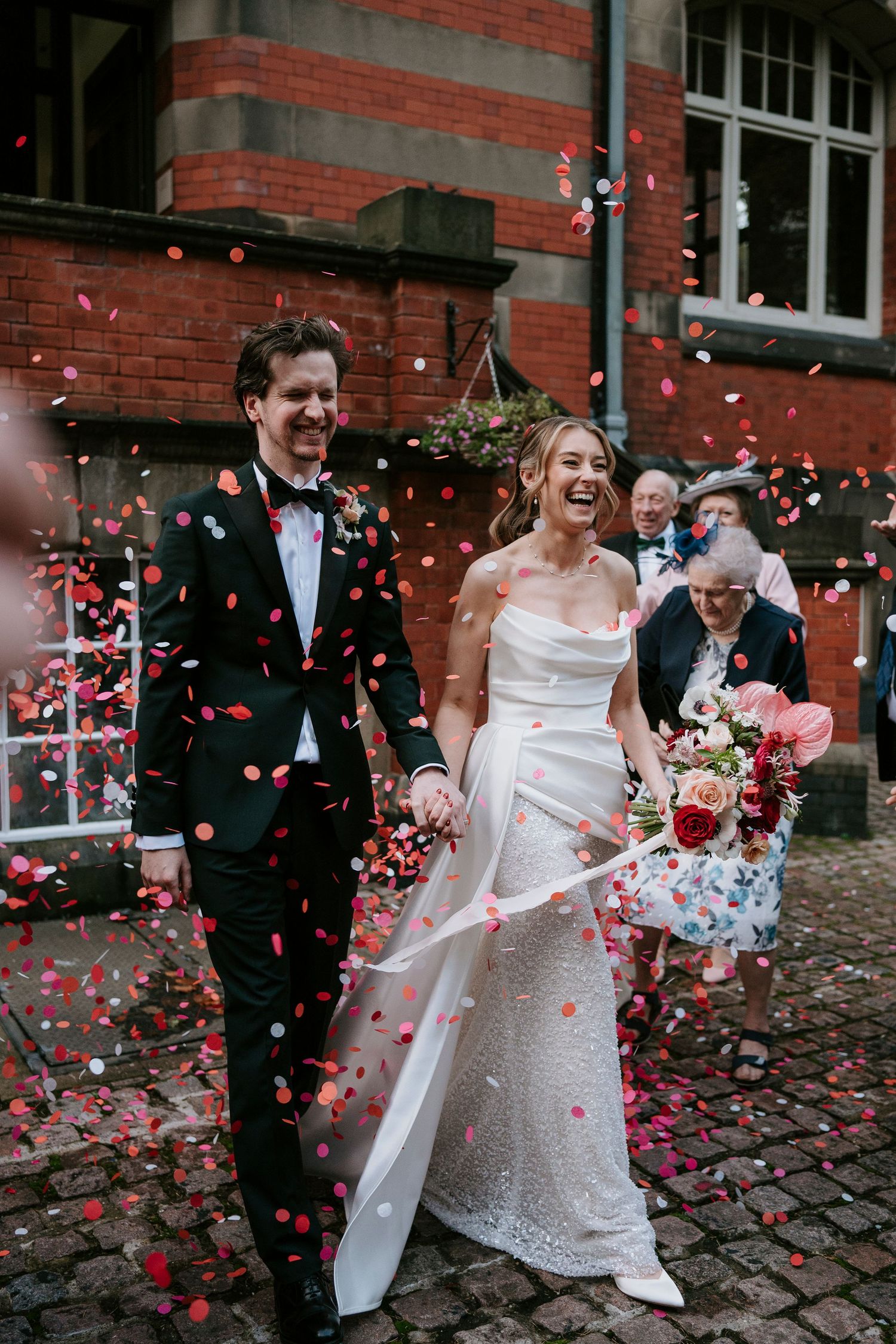 Wedding couple celebrates under a shower of pink confetti outside a brick building.