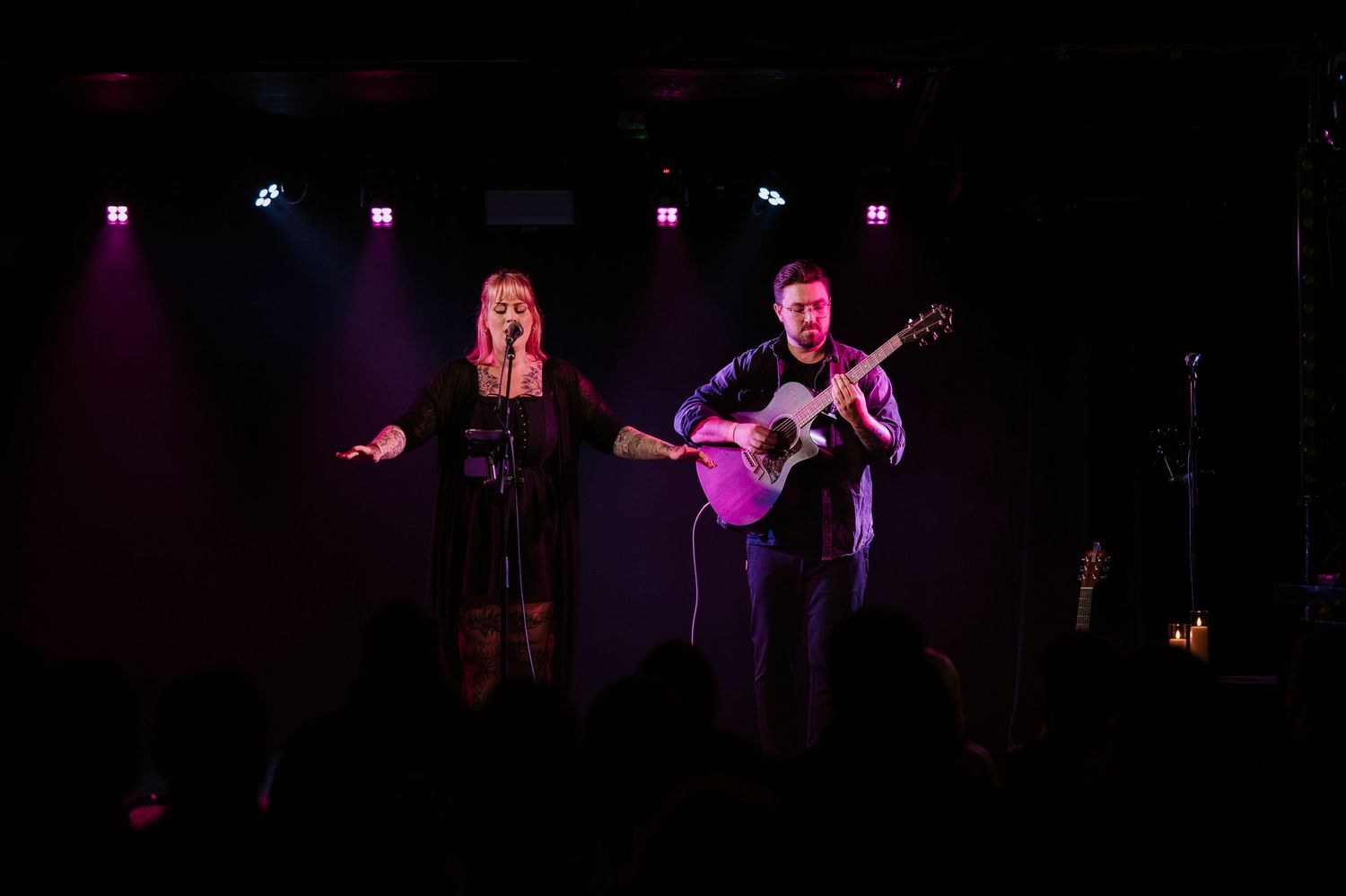 Musicians perform on a dimly lit stage with guitars and dramatic purple lighting creating an atmospheric concert scene.
