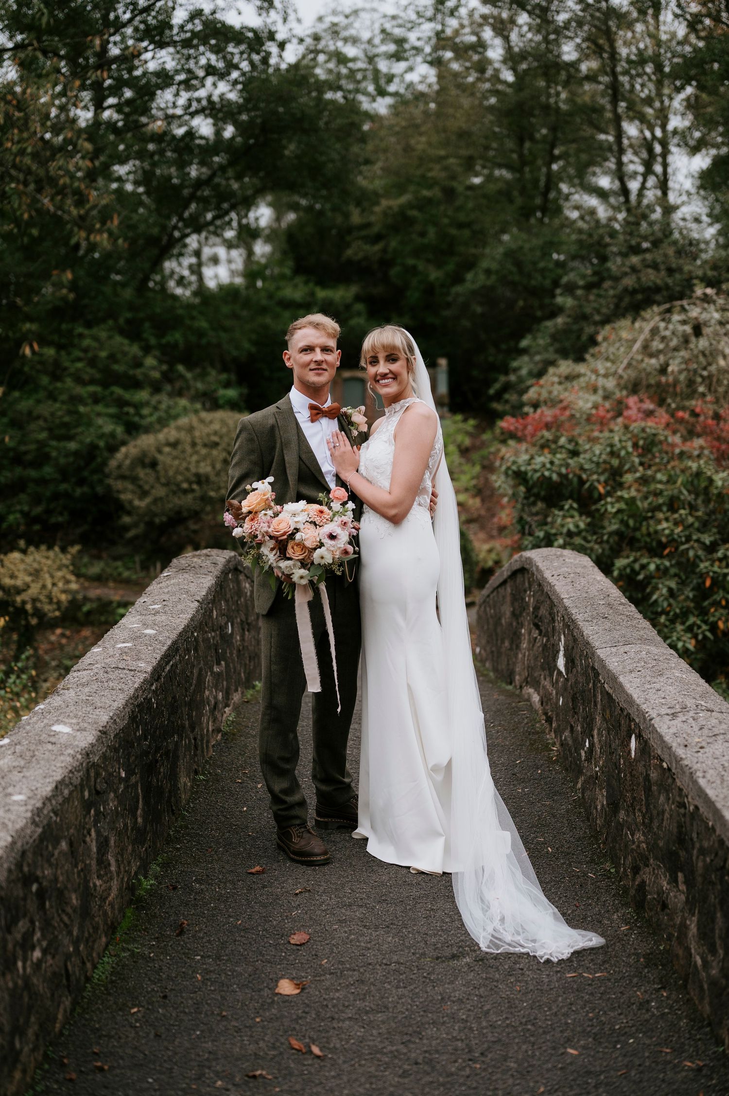 A couple stands together on a stone bridge surrounded by lush greenery during an autumn wedding ceremony.