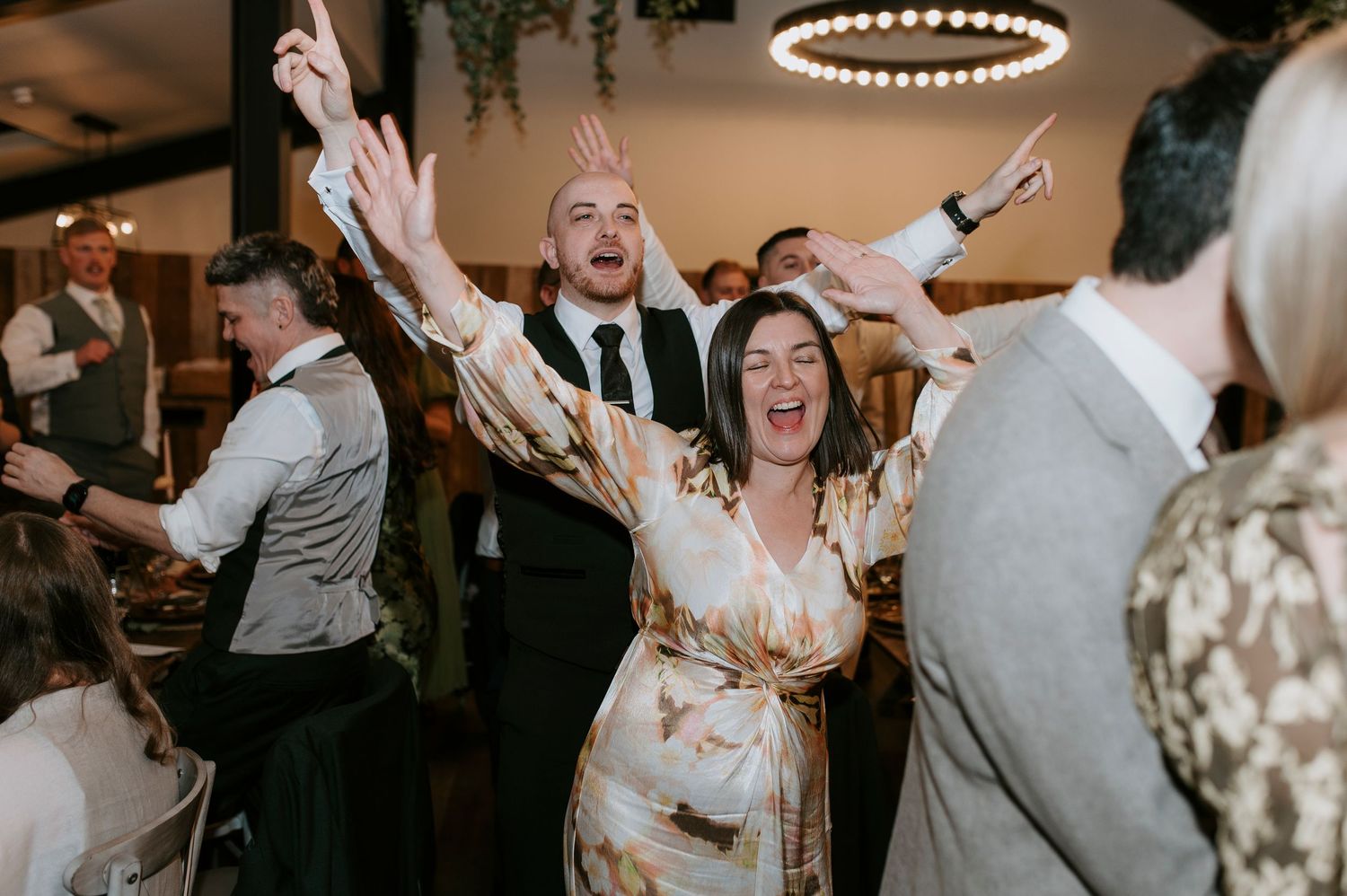 Wedding guests dance and celebrate enthusiastically on a dimly lit dance floor at an indoor reception.