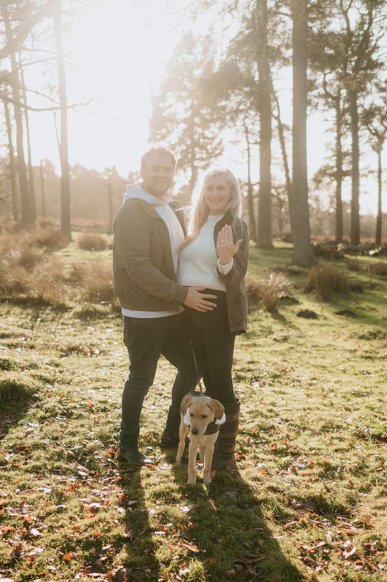 A couple and their dog enjoy a romantic autumn photoshoot in a sunlit forest with fallen leaves.