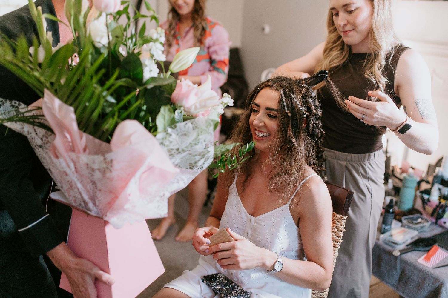 A group of people gather together in a bright room with pink floral bouquets and celebratory atmosphere.