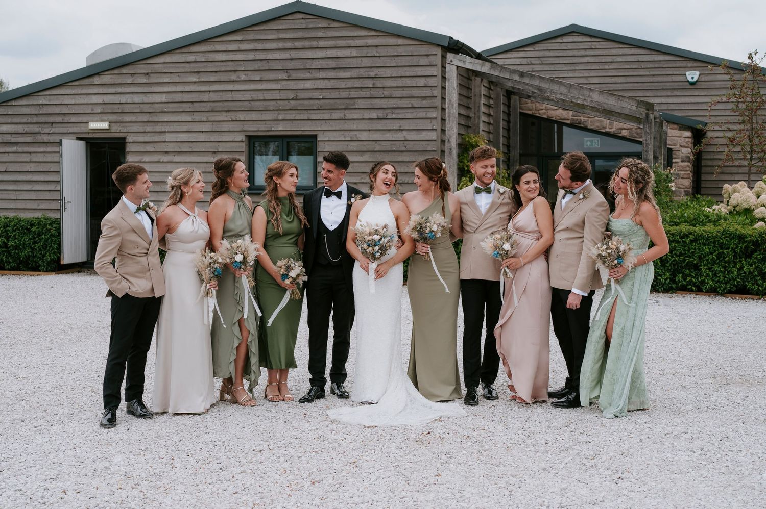 Wedding party in pastel dresses and suits poses together on white gravel path outside rustic wooden buildings.