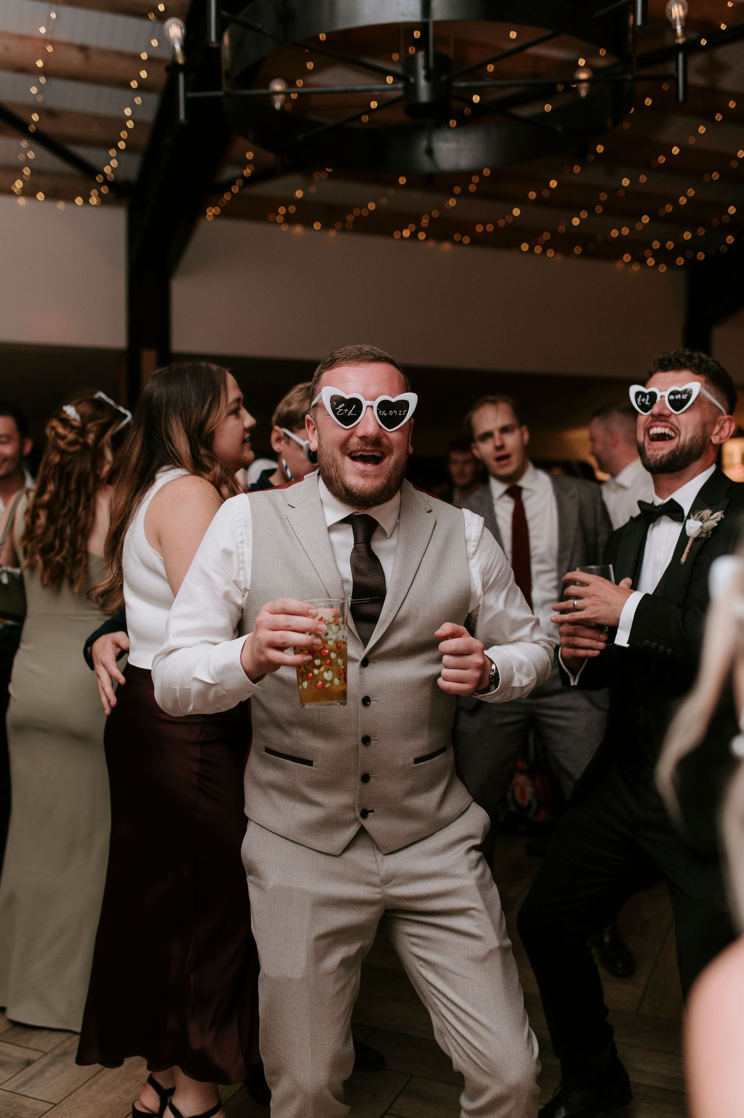 Wedding guests in gray vests dance and celebrate at a reception under twinkling string lights.