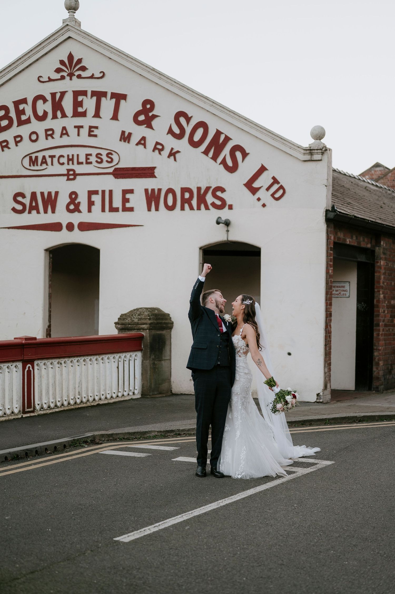 Joyful newlyweds celebrate outside Beckett & Sons Saw & File Works, groom raising fist while bride holds bouquet.