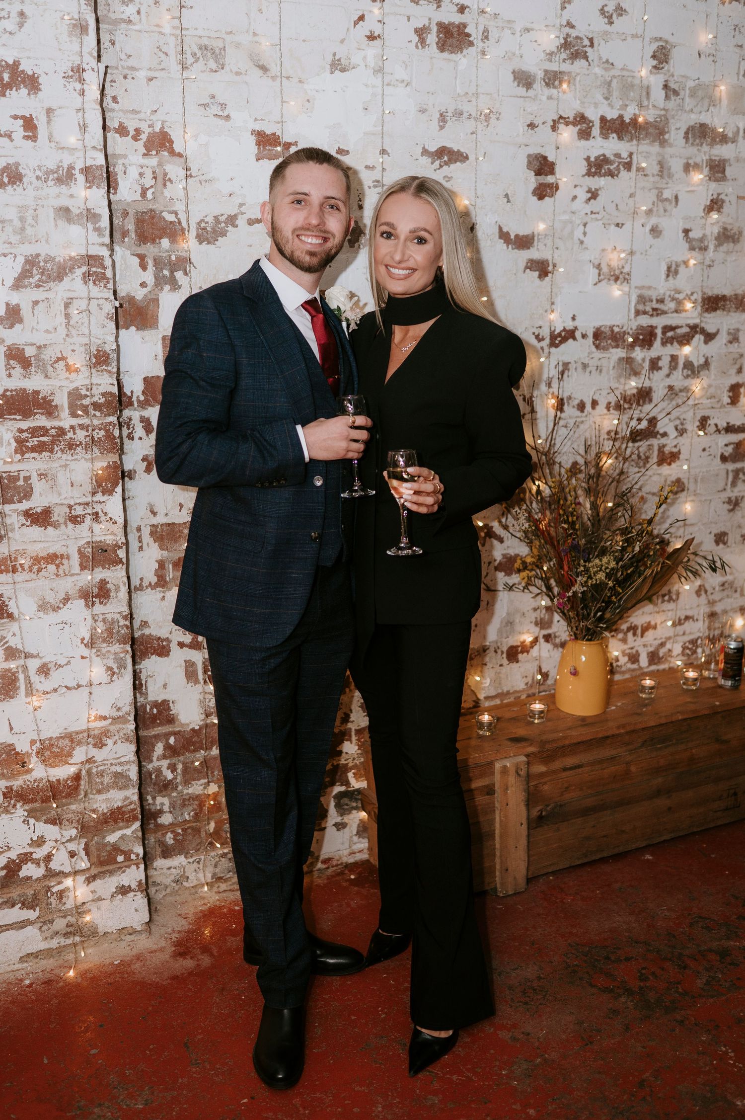 Couple in formal attire holding champagne glasses posing in front of a rustic brick wall with fairy lights.