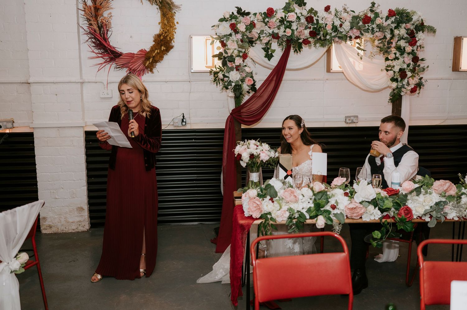 Wedding ceremony with officiant reading at a floral-decorated head table in an industrial venue.