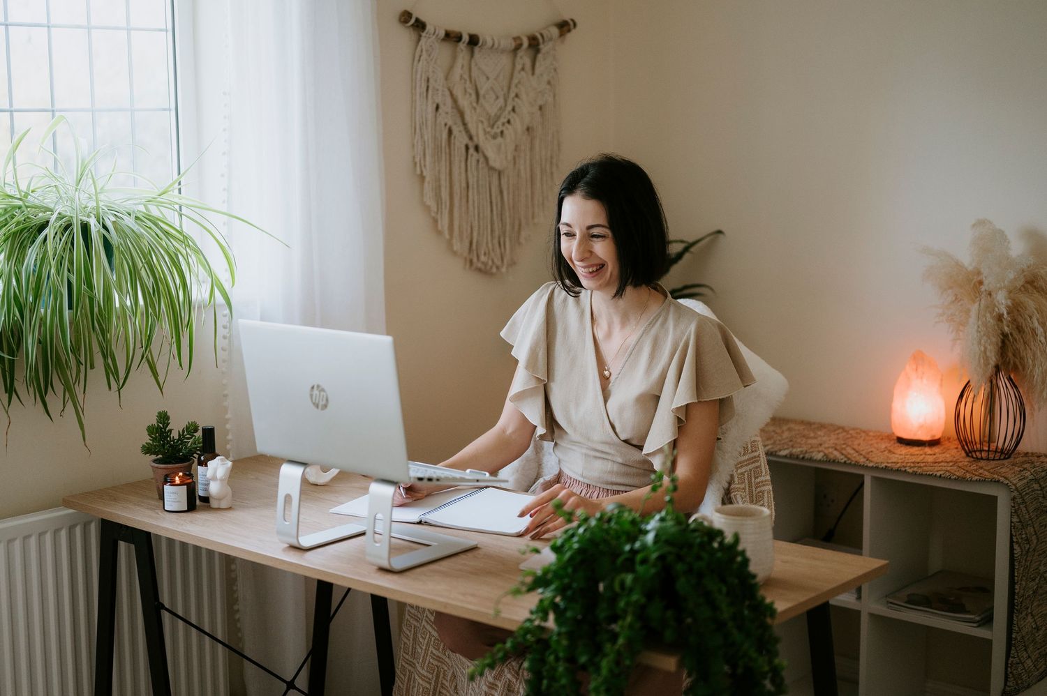 Woman working at minimalist home office desk with plants, macrame wall decor, and warm lighting.