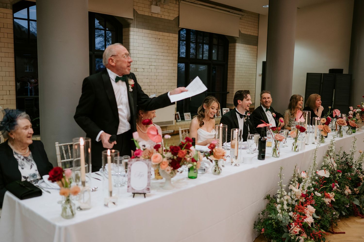 Wedding guests seated at a long outdoor reception table decorated with floral centerpieces and wine glasses.