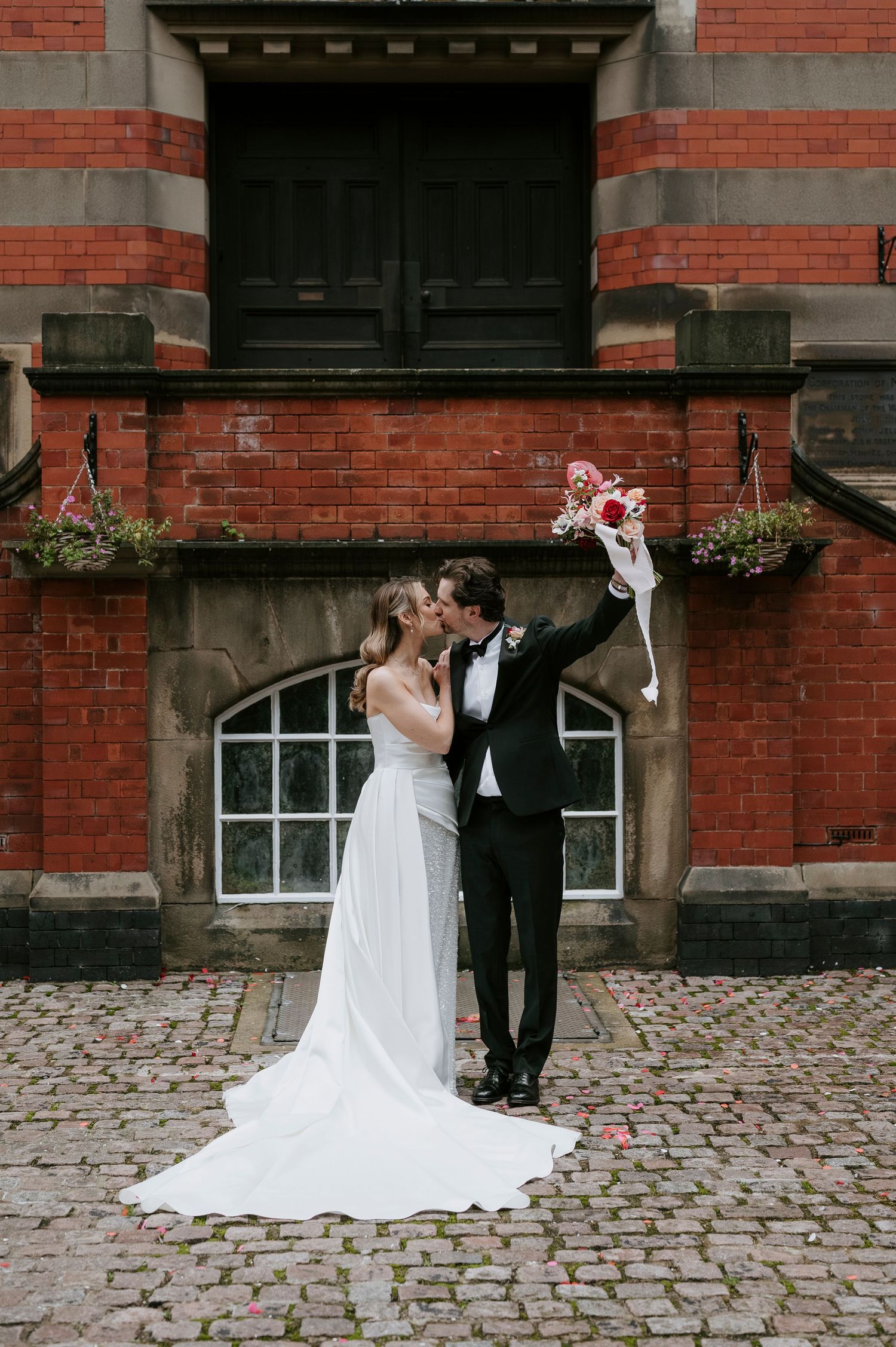 A romantic wedding couple shares intimate moments against a rustic brick building backdrop with cobblestone pavement.