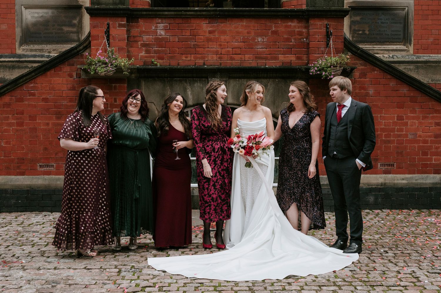 Wedding party gathers on cobblestone path in front of historic red brick building.