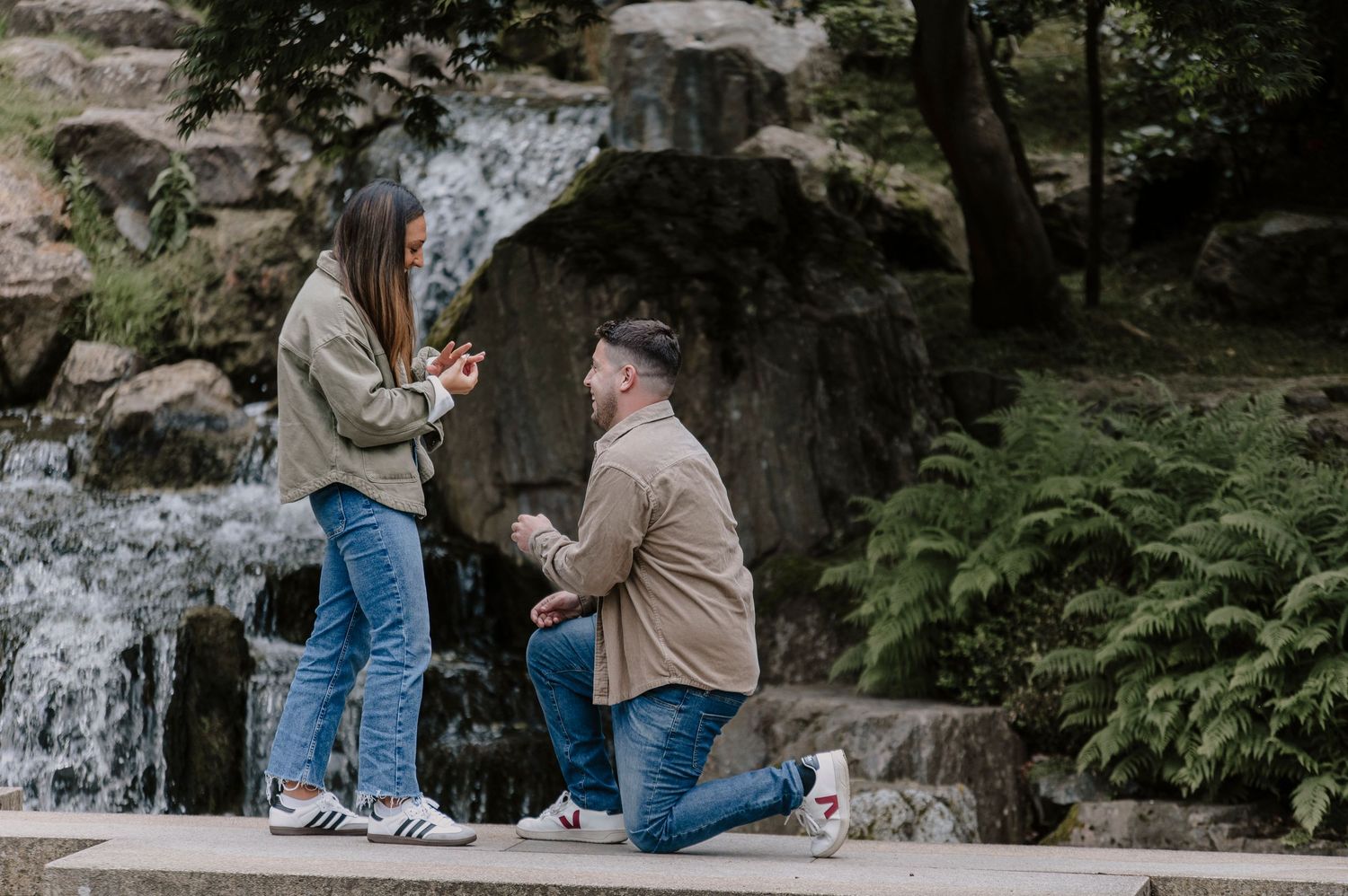 A couple in casual attire kneels on rocks near a waterfall during an outdoor engagement photo session.
